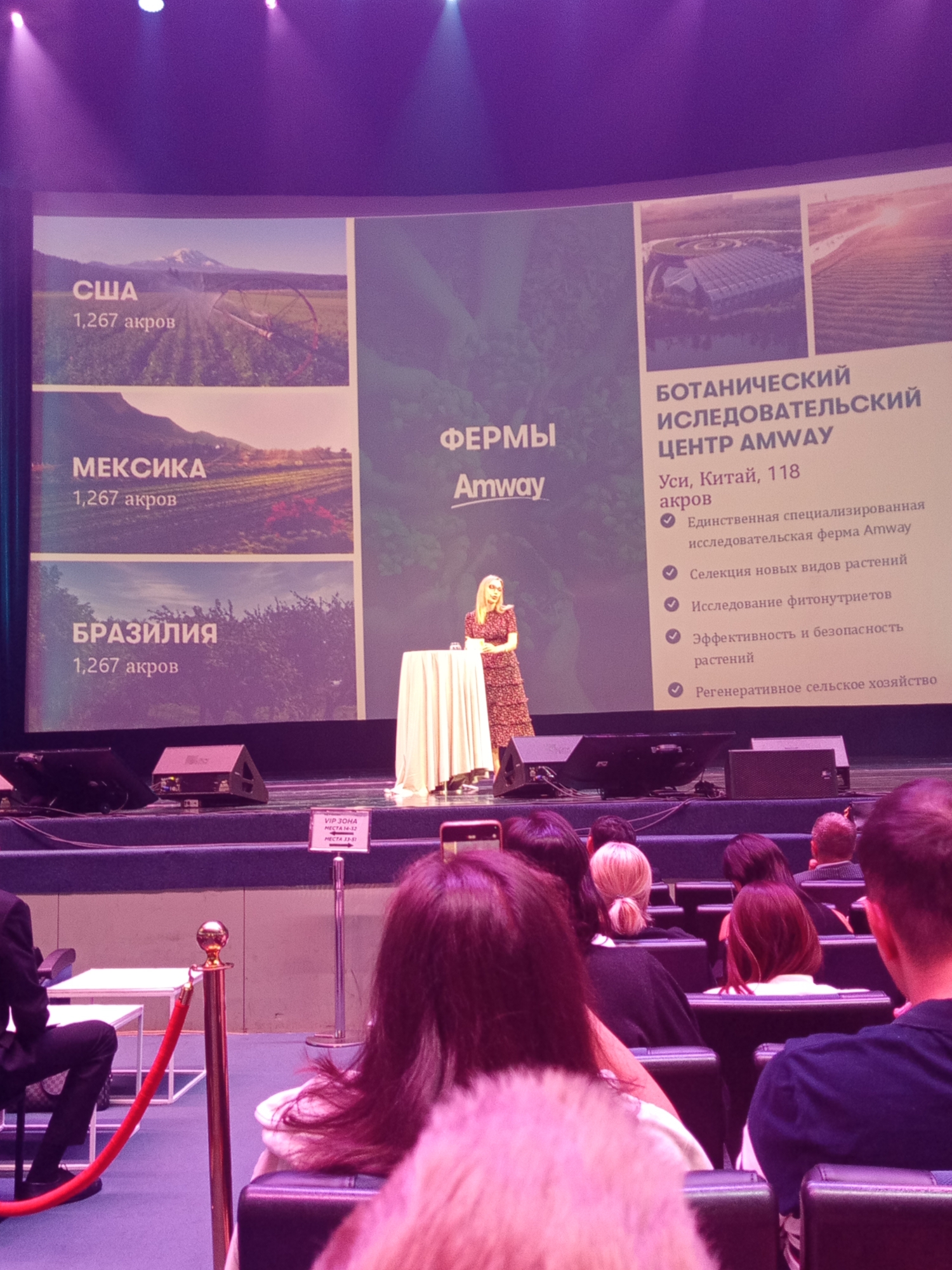 The image shows a presentation taking place in a venue, likely a conference hall or auditorium. A large screen displays information about Amway farms and a botanical research center. Text on the screen is in Russian. The screen is divided into sections, with images of farms in the USA, Mexico, and Brazil. To the right of these images, there is a section titled "BOTANICAL RESEARCH CENTER AMWAY" with details about its location in Wuxi, China, and its key features.
In front of the screen, a woman in a dress stands next to a table covered with a white tablecloth, seemingly giving a presentation. The audience is seated in the foreground, with many people visible, their backs to the camera. The lighting appears to be stage lighting, with some purple hues visible in the background. There are also red ropes and stanchions in the foreground, suggesting a VIP or restricted area. In the bottom right corner, a sign in Russian reads "VIP ZONE SEATS 14-32 SEATS 33-51" with an arrow pointing to the right.
The presence of Russian text on the screen and the sign, combined with the general ambiance of a large-scale presentation, strongly suggests that this event is taking place in Saint Petersburg, Russia. The venue appears to be well-equipped for presentations, with a large screen, stage, and audience seating.