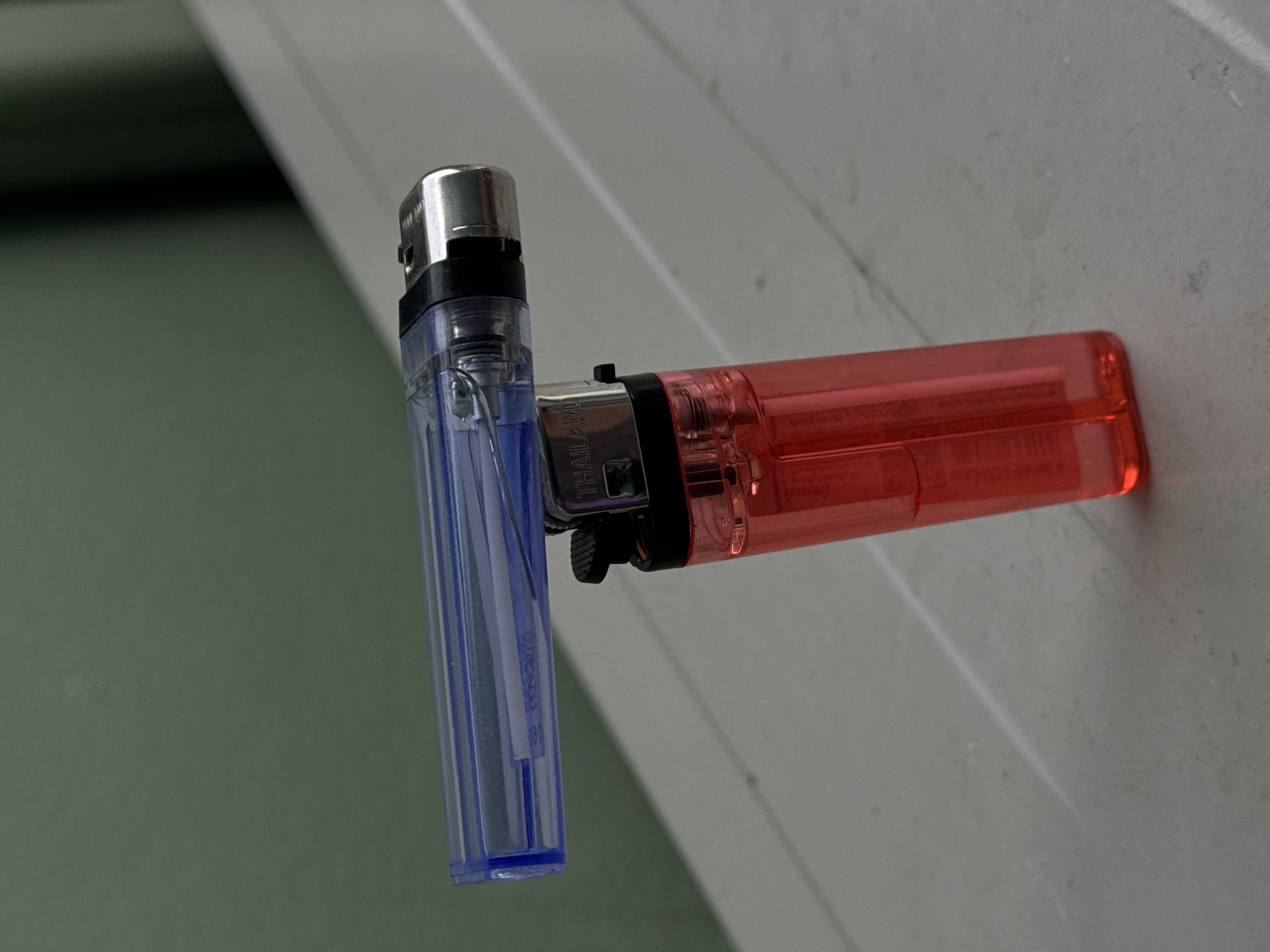 Two lighters are positioned on a white surface, likely a table. A red lighter stands upright, while a translucent blue lighter rests horizontally on top of it, its nozzle intersecting the red lighter's body. The background is a plain, muted green wall. The scene appears to be indoors, possibly in a residential or commercial setting within Kathu, Thailand. There are no individuals or animals visible in the image.