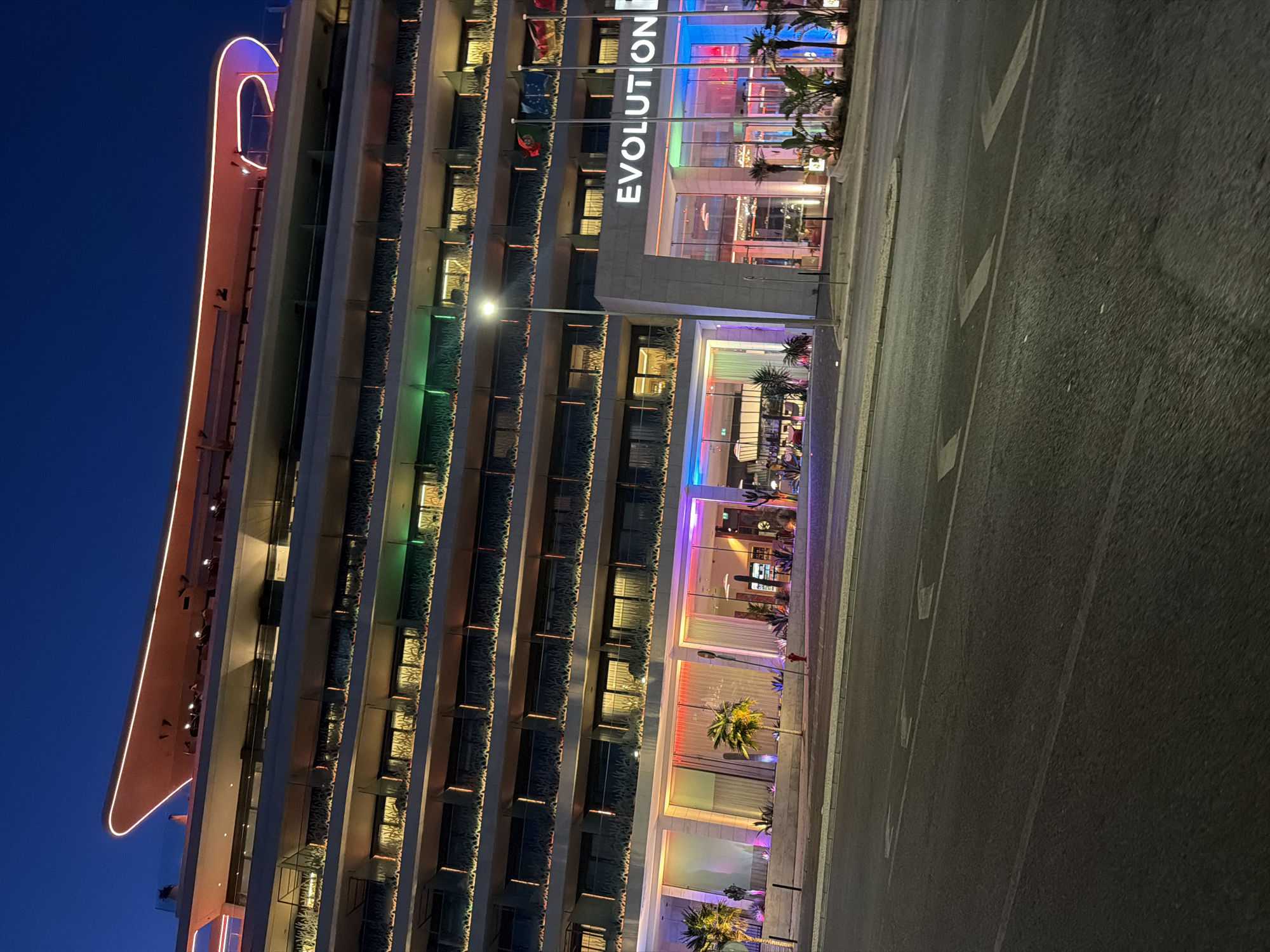 The image shows the exterior of the Evolution Hotel in Cascais, Portugal, at dusk. The hotel is a modern, multi-story building with a curved, illuminated roofline. The facade is lit with colorful lights, particularly around the ground floor entrance and balconies. Several flags are visible on poles outside the entrance. Palm trees and other tropical plants are planted in front of the hotel, and a street with lane markings runs in the foreground. The sky is a deep blue, indicating it is twilight. There are no people clearly visible in the image.