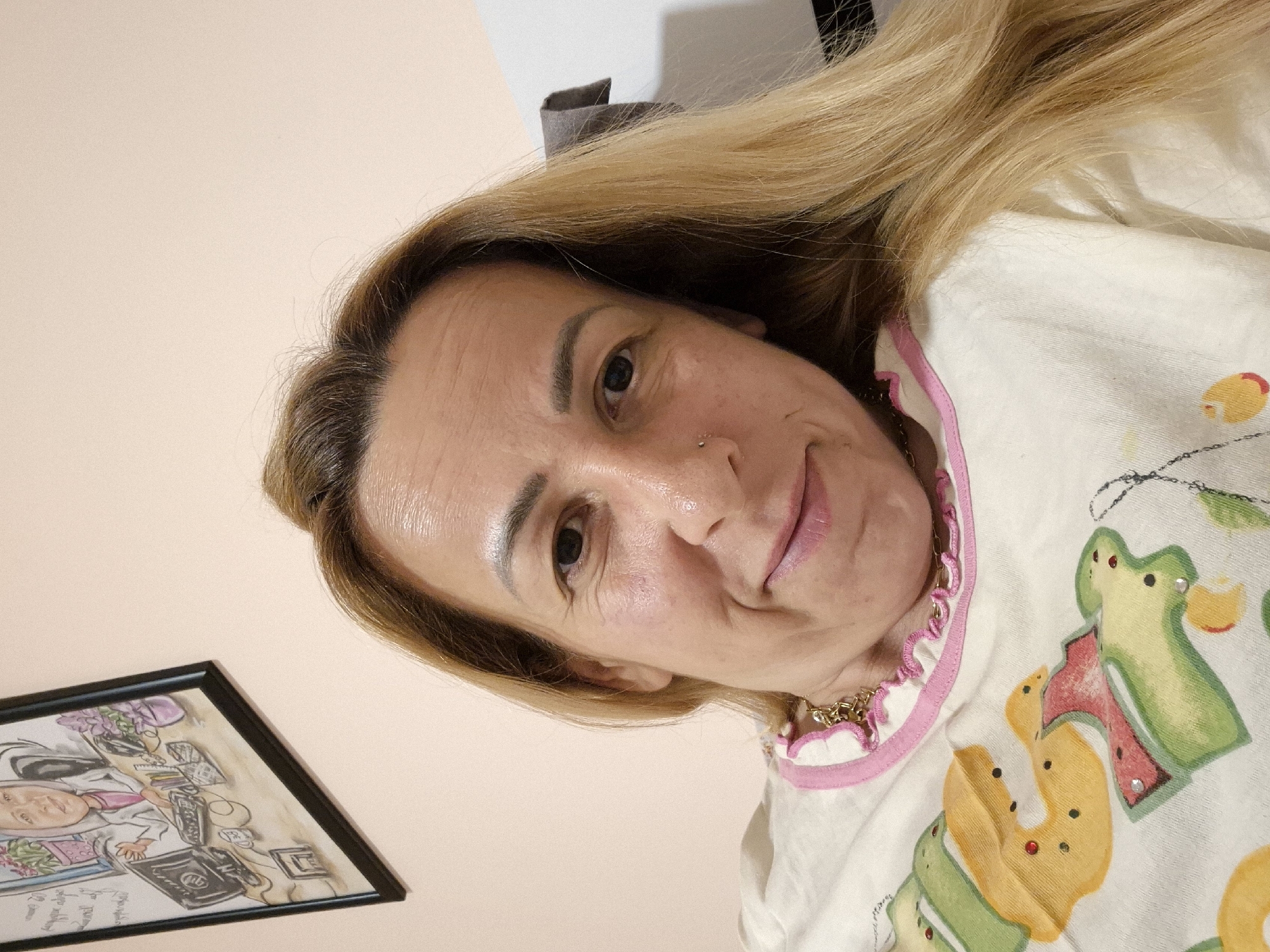 A medium shot is provided of a woman with blonde hair and light-colored skin. She is looking directly into the camera and smiling. The woman is wearing a white shirt with a pink ruffled collar and a gold chain necklace. Behind her is a light-colored wall, and to her left, a framed drawing of a person sitting at a desk is visible. The drawing depicts a person with a headscarf, sitting in front of a computer with plants and other office supplies on the desk. The background also shows a part of a white piece of furniture and a gray fabric hanging from it.