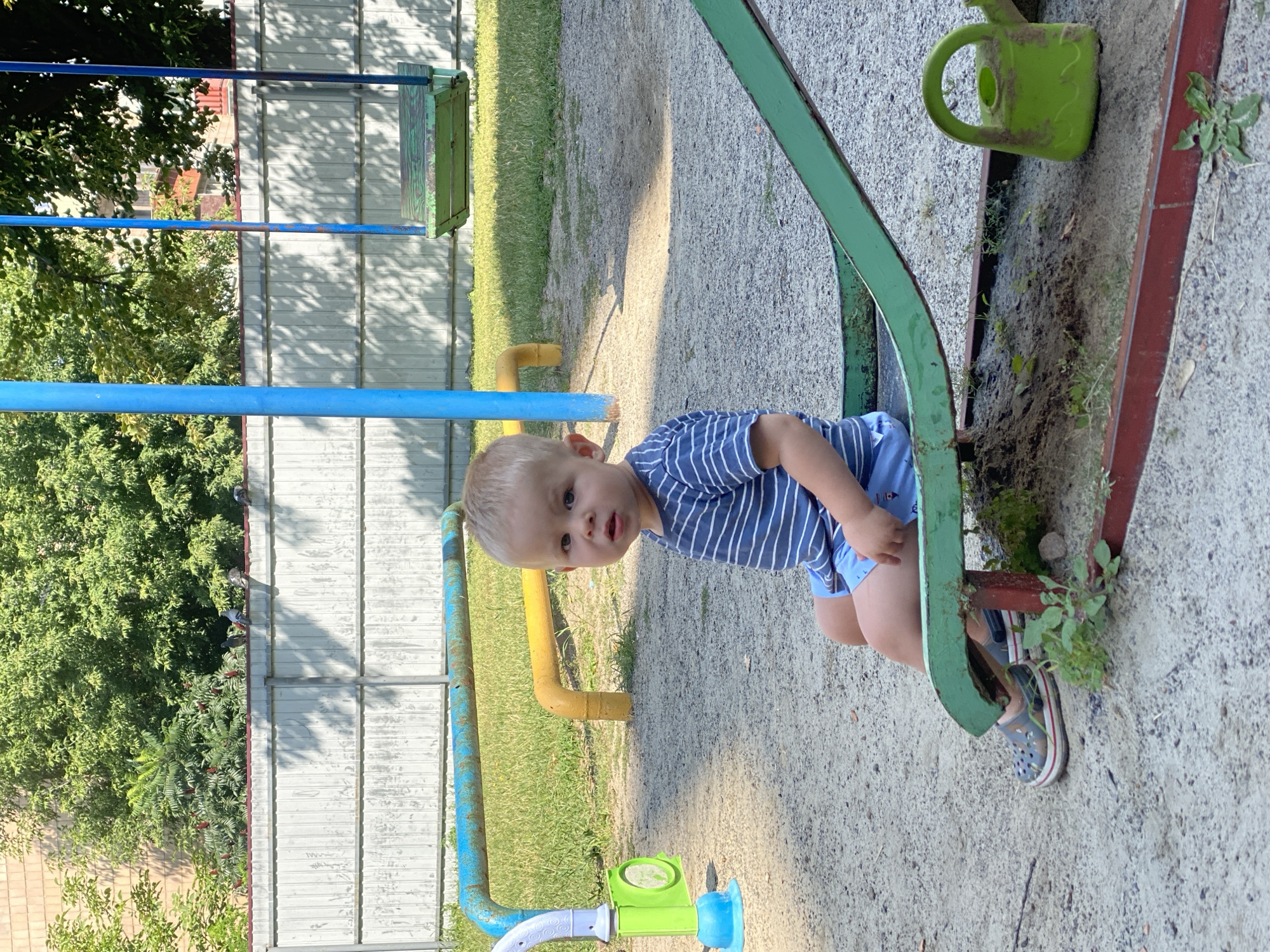 A light-skinned toddler with blond hair sits on a green metal slide at a playground. The child is wearing a blue and white striped t-shirt and blue shorts. In the background, there is a swing set with a green seat, a blue pole, and a metal fence. Trees and buildings are visible behind the fence. A green watering can is on the ground next to the slide. The ground is covered with sand and gravel. The image appears to be taken on a sunny day in Poltava, Ukraine.