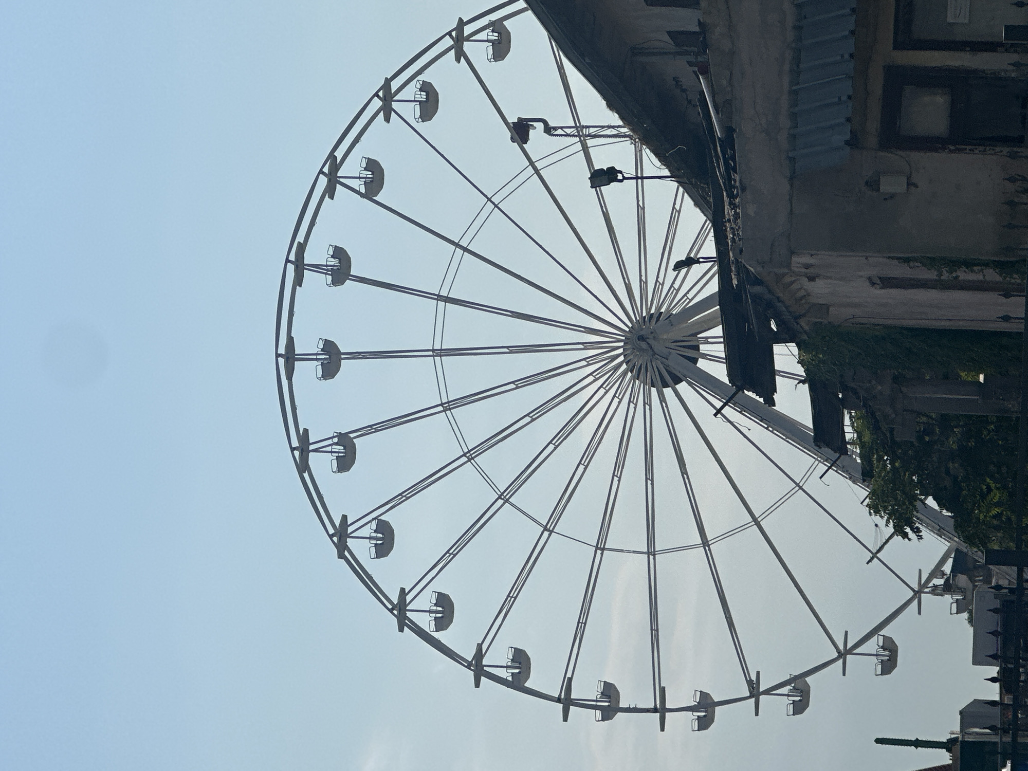 A Ferris wheel is positioned against a clear, pale blue sky, with only the top half of the structure visible. The Ferris wheel is white and appears to be made of metal, with numerous spokes radiating from the center. Several empty gondolas, also white, are suspended from the outer rim of the wheel. To the right of the Ferris wheel, the roofline of a weathered, old building is visible, its dark, textured surface contrasting with the bright sky. Part of a dark green, leafy plant is growing on the side of the building. The lower part of the image shows a dark metal fence and some indistinct structures, suggesting an urban environment.
