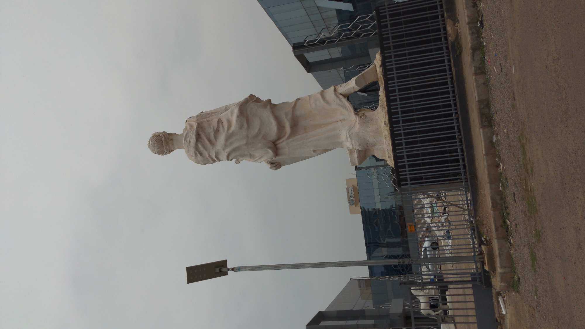 A large, light-colored statue of a robed figure, possibly male, stands prominently in the mid-ground, facing away from the viewer and slightly to the left. The figure is draped in flowing garments, with details visible in the fabric folds and the hairstyle. The statue is placed on a simple base.
In the background, several modern buildings with glass facades are visible, reflecting the overcast sky. A tall pole, topped with a rectangular light fixture, possibly a solar panel or street light, stands to the left of the statue.
The foreground features a dark metal fence or gate running horizontally across the lower right portion of the image. Beyond this fence is an unpaved, dusty road or dirt track, with some sparse green vegetation along its edge. The sky is uniformly overcast, indicating a cloudy day with diffused natural light.
No specific activity or event is taking place; the scene is static. On one of the buildings in the distance, a small sign is visible, but any text on it is too indistinct to read clearly, though it appears to feature characters that could be Arabic script or a similar language. The setting suggests an urban or commercial area, possibly a public space or the entrance to a business complex.