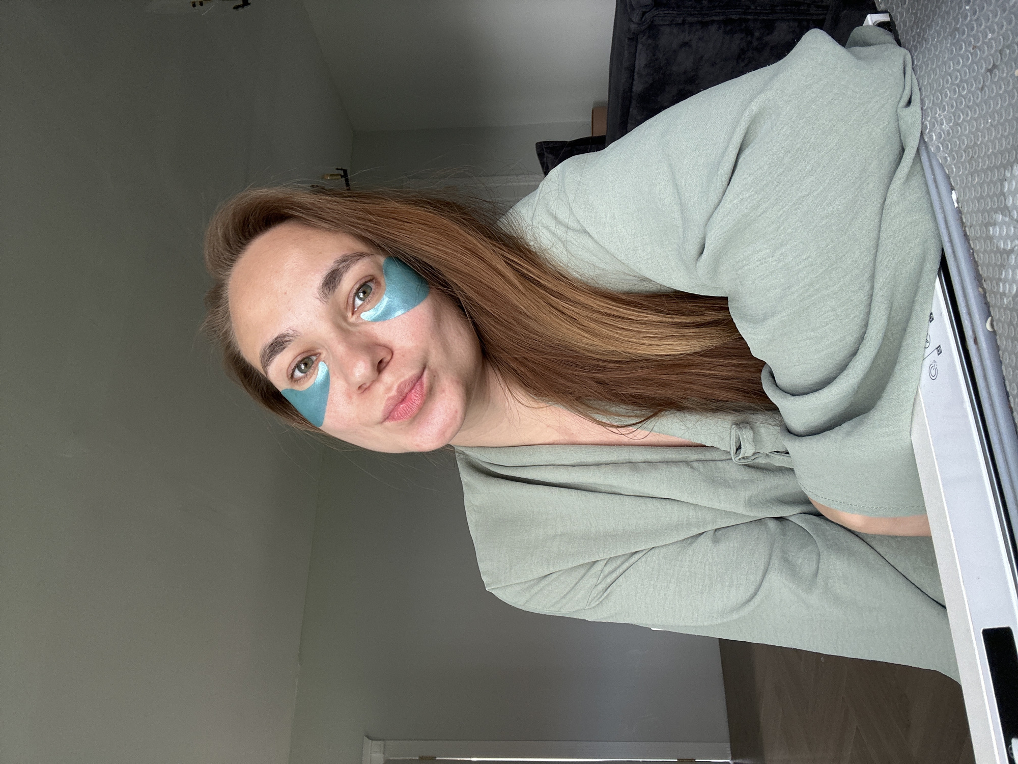 In Kharnavy, Russia, a young woman with long, auburn hair sits indoors, looking directly at the camera. She is wearing a light green, textured robe. Blue under-eye patches are visible on her face. Her lips are pursed. The background consists of a plain wall and a dark sofa. A white surface, possibly a desk or table, is in the foreground. The lighting is soft and natural.