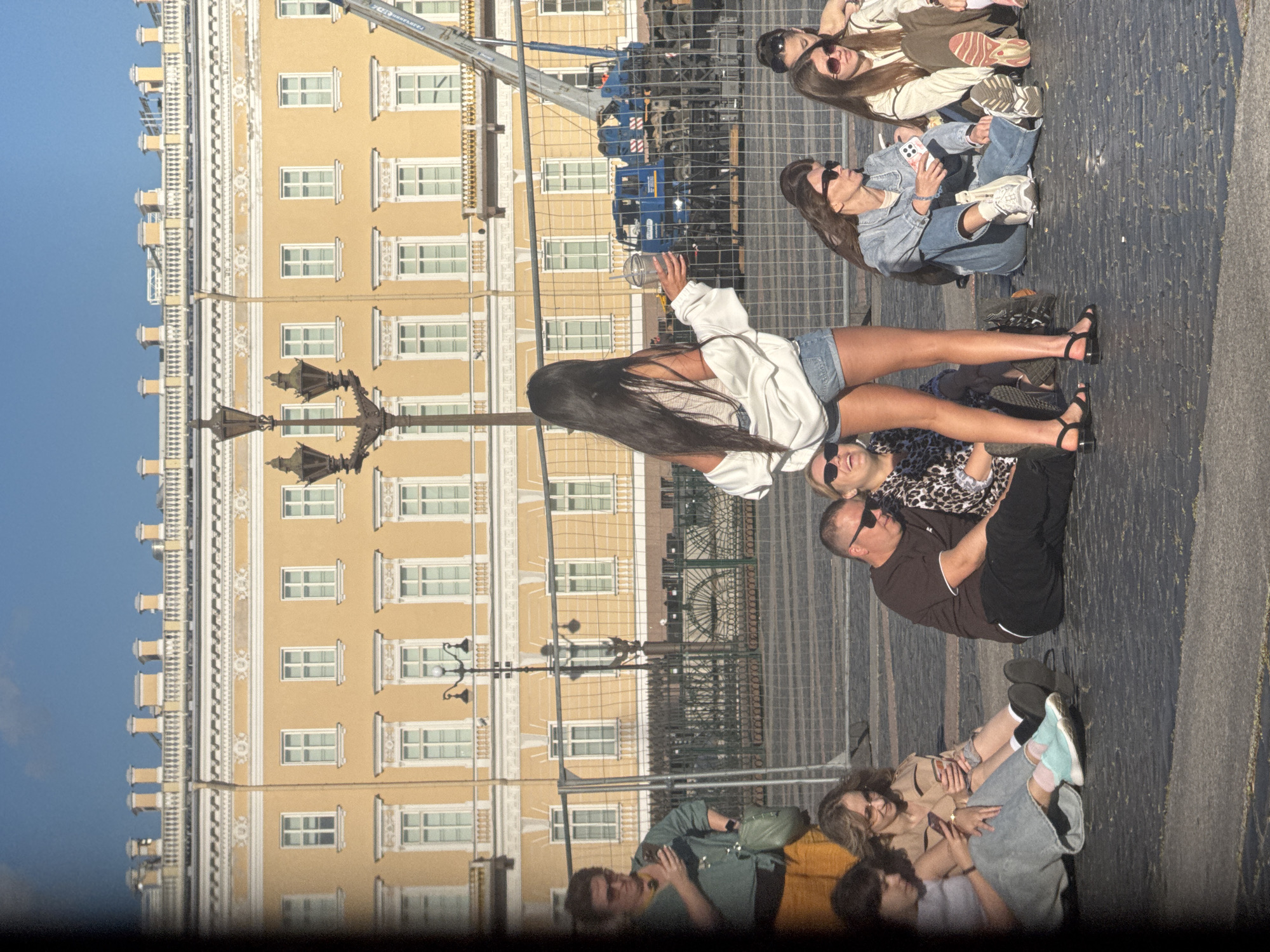 The image captures a sunlit scene in front of a grand, yellow neoclassical building, likely a palace or government building, in Saint Petersburg, Russia, given its architectural style. The building features multiple stories with evenly spaced windows, ornate white trim, and a decorative balustrade along the roofline. In the foreground, a group of people are seated on the cobblestone pavement. A woman with long dark hair, wearing a white open shirt and denim shorts, stands facing away from the camera, holding a clear plastic cup with a straw. To her left, a woman in a green shirt and yellow pants is looking at her phone. Other individuals are seated in a relaxed posture, some using their phones, others conversing. Behind the seated group, a temporary fence separates them from a construction area where a large blue vehicle, possibly a truck or crane, is visible, along with a taller construction crane extending upwards. The sky is clear and blue. The overall atmosphere suggests a casual gathering in a public space on a pleasant day.