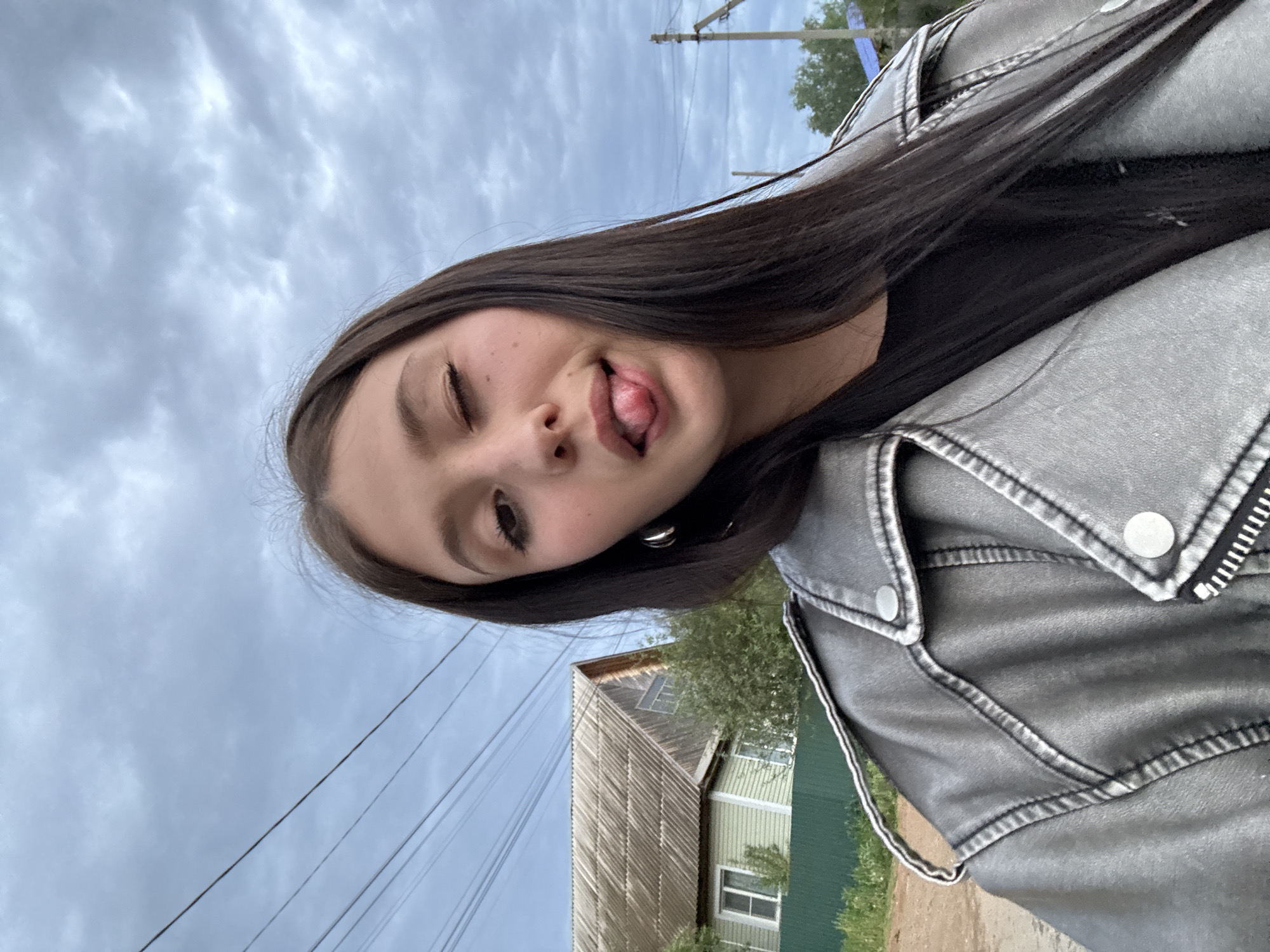 A woman with dark hair and wearing a gray leather jacket is sticking out her tongue and winking. She is outdoors, with a house visible in the background. Overhead power lines are also visible, and the sky is overcast. The photograph appears to have been taken at an angle. It is not possible to ascertain whether the location is Фоки, Russia, or to verify that information from the image.