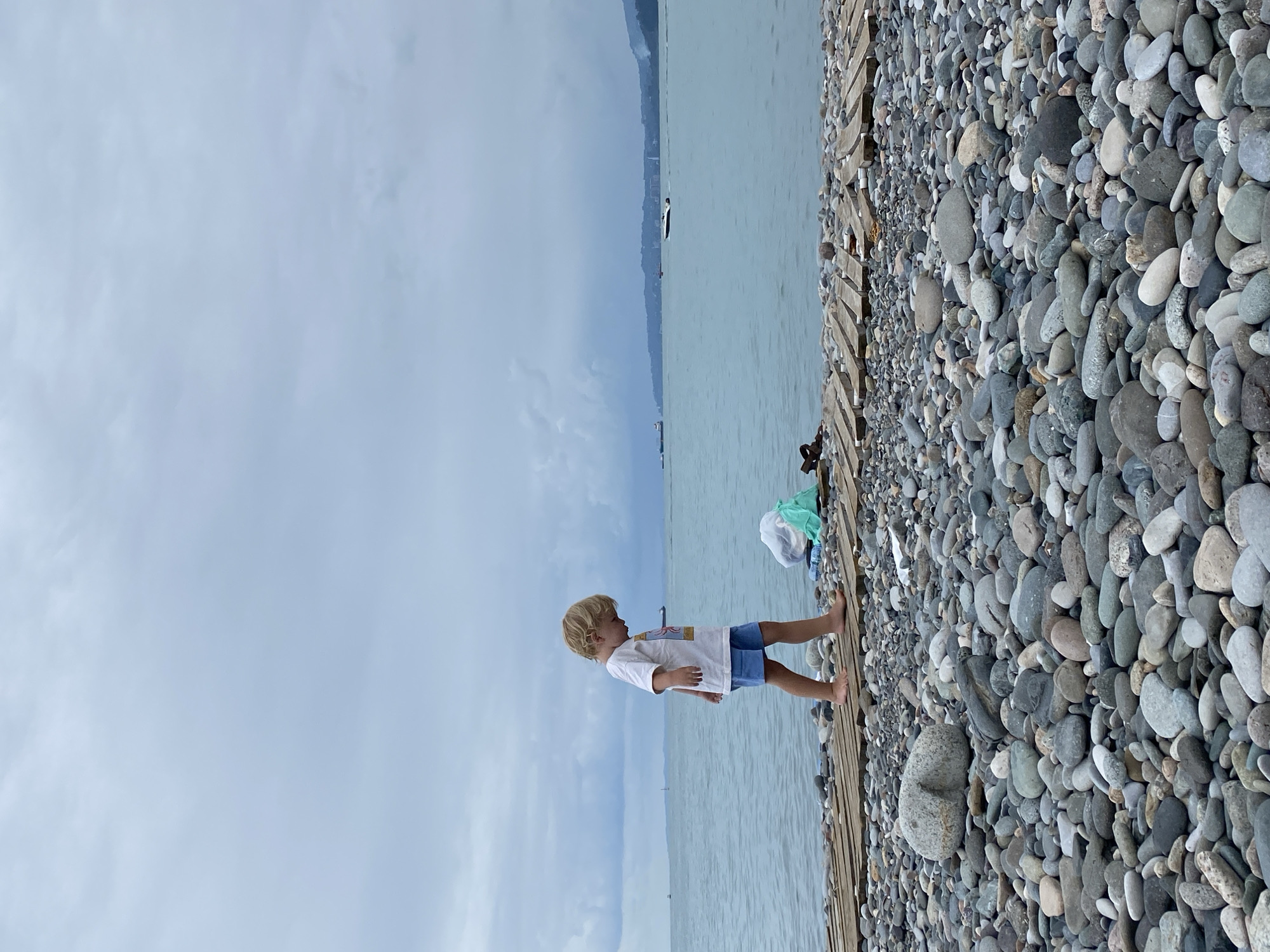A young boy with blond hair is walking on a wooden pier on a pebble beach in Batumi, Georgia. He is wearing a white t-shirt with a graphic print and blue shorts. The boy is looking down as he walks. To his right, a green plastic bag and a white plastic bag are lying on the pier. The sea is calm with a few boats visible in the distance. The sky is overcast with dark clouds. The background shows a coastline with trees and buildings. The overall scene depicts a quiet and somewhat somber atmosphere due to the cloudy weather.