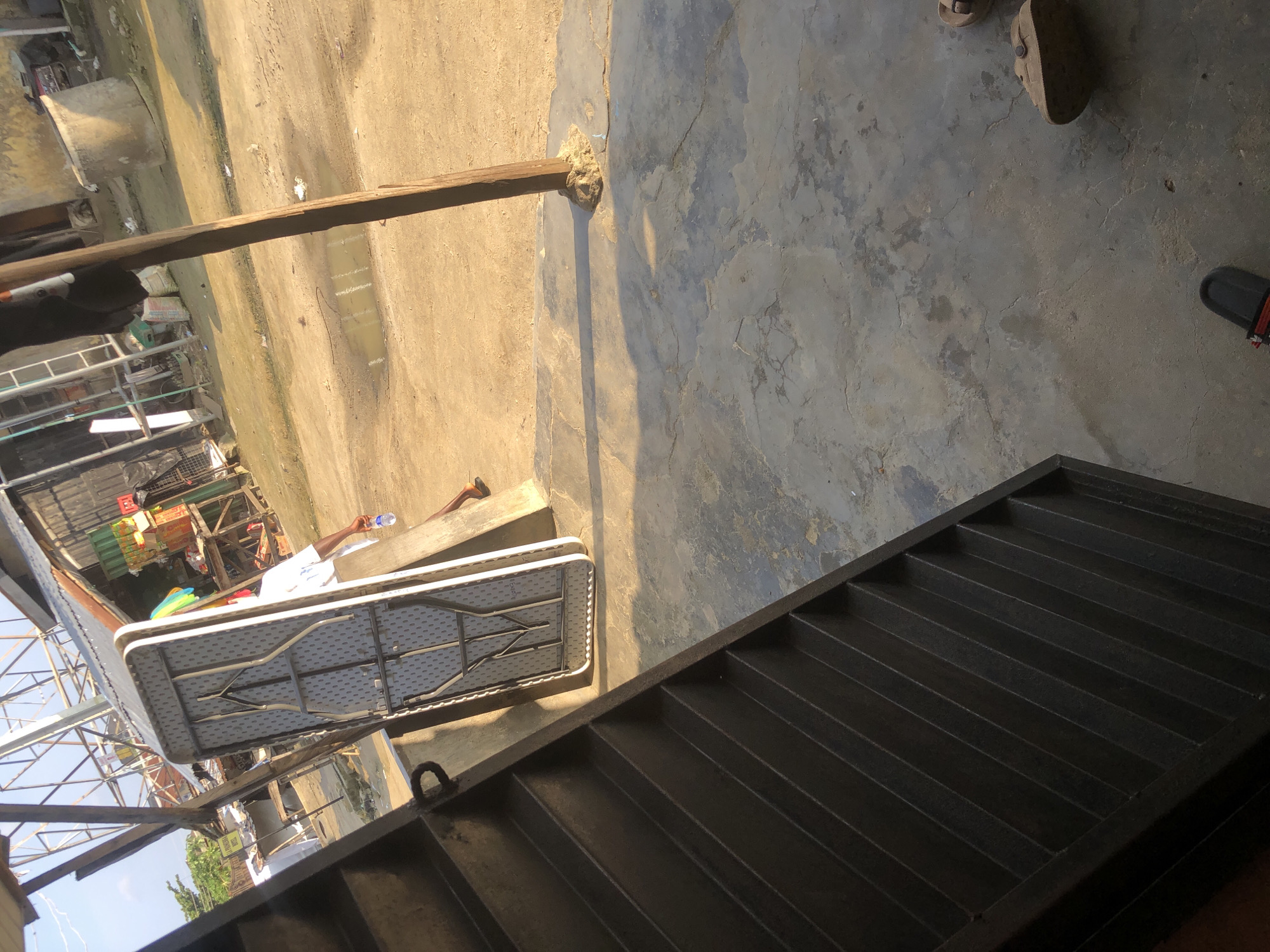 In Ado, Nigeria, a folding table is propped against a concrete wall near a dirt path. A person wearing a white shirt and shorts walks away from the table, holding a bottle of water. In the foreground, a dark metal staircase descends to the left. The background shows a market area with stalls and structures, including what appears to be a cement mixer and various goods. The ground is a mix of dirt and concrete, with some puddles of water visible. A wooden post stands vertically in the mid-ground. Two pairs of sandals are visible on the concrete surface in the lower right of the image.