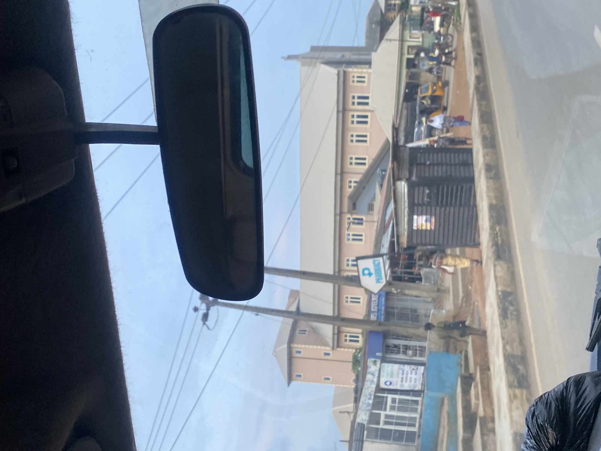 The image depicts a street scene in Ikorodu, Nigeria, as viewed from inside a vehicle. A dark rear-view mirror is prominently positioned in the upper center of the frame. The foreground shows a portion of the vehicle's interior, including a black trash bag on the left and part of the dashboard with what appears to be windshield wipers.
The middle ground of the image reveals a street with a sidewalk and a road. On the sidewalk, several individuals are walking. One person in a dark shirt and trousers walks away from the camera, while another in a yellow dress walks towards the camera. Several buildings line the street. One building, a two-story structure with a light brown facade and a prominent cream-colored roof, has multiple windows. Adjacent to it is a multi-story building with a similar cream-colored roof, featuring a steeple-like structure, suggesting it might be a church. Signs are visible on some of the buildings, including one that reads "PHARMACY" with a blue symbol of a woman. There are also smaller structures and market stalls along the sidewalk.
In the background, several power lines are visible against a cloudy, light blue sky. A yellow auto-rickshaw, commonly known as a Keke Napep, is parked on the street, and several motorcycles are also present. The overall environment suggests a commercial or residential area within a Nigerian city. The lighting indicates it is daytime, possibly overcast or late afternoon.