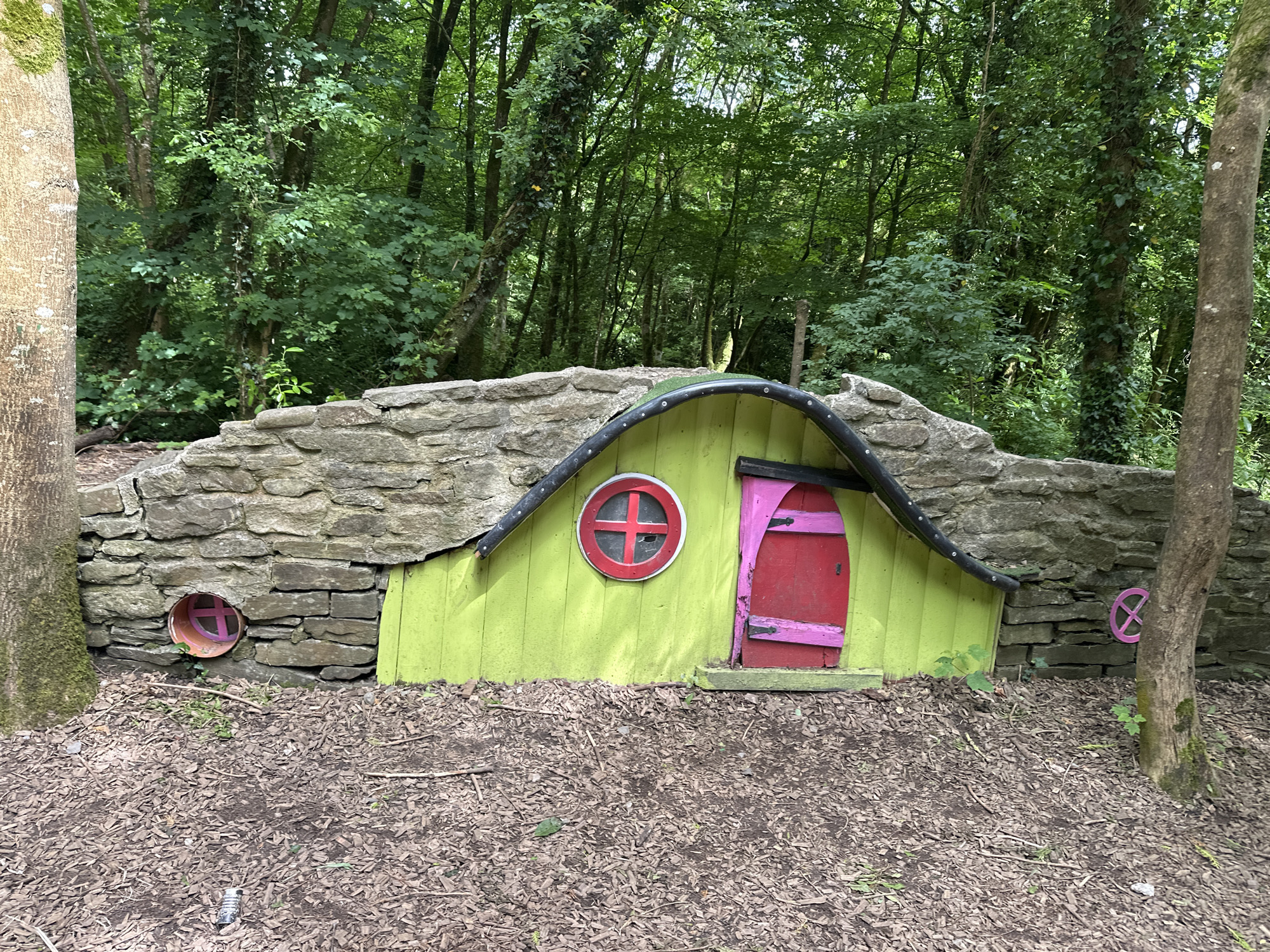 The image depicts a whimsical, fairy-tale-like structure nestled within a woodland setting, likely located in the townland of Cretten Commons, Castlecomer, Ireland. The structure is partially built into a low stone wall, with a brightly colored, lime-green facade. A curved, black-rimmed roof overhangs the facade.
The facade features a vibrant pink, arched door with visible hinges and a small latch. To the left of the door, a circular window with a red frame and a cross-shaped design inside is visible. To the left of this window, further into the stone wall, is a smaller circular opening with a pink cross-shaped design. To the right of the main structure, a pink, wheel-like decorative element is affixed to the stone wall.
The ground in front of the structure is covered with wood chips and fallen leaves, with scattered twigs and small green plants emerging from the mulch. The background is dominated by a dense forest with tall trees and lush green foliage, indicating a natural, outdoor environment. Two tree trunks are prominently visible on the left and right edges of the frame, one on the left appearing thicker and moss-covered. The lighting suggests an overcast or shaded day, with soft, diffused light. No people are present in the image.