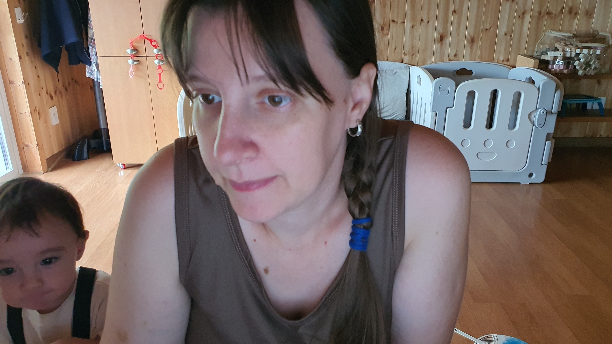 Two individuals, an adult and a toddler, are captured in a close-up, indoor shot. The adult, positioned closer to the camera and taking up the right half of the frame, has dark hair pulled back into a braid secured with a blue hair tie. They are wearing a brown tank top. To the left and slightly further back, the toddler, facing forward, has dark hair and is wearing a white shirt with black suspenders.
The background reveals a room with light-colored wooden walls and a polished wooden floor. To the left, a wooden cabinet with drawers and doors is partially visible, with a string of red and silver decorative items hanging from it. Behind the adult and to the right, a light gray plastic playpen with circular and slotted openings is situated. In the upper right corner, a shelf displays several boxes of what appear to be wooden toys or building blocks. The lighting suggests an indoor setting, possibly a home environment, with natural light sources not explicitly visible but contributing to the overall illumination.