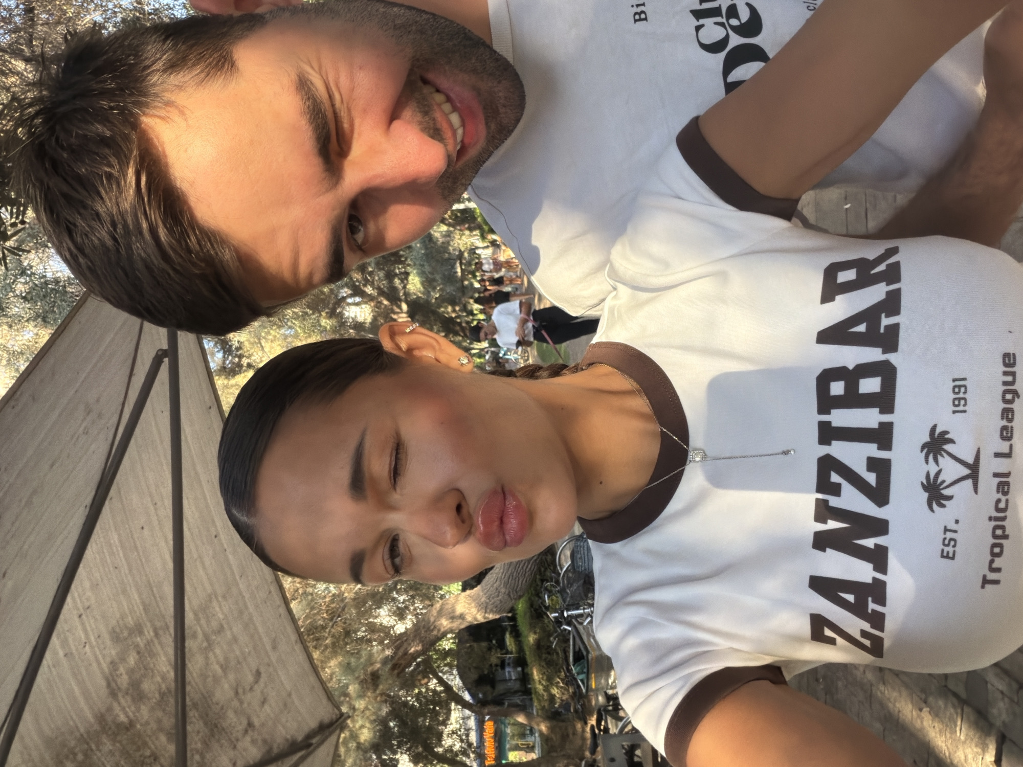 A candid, close-up selfie shows a man and a woman standing outdoors, possibly in Tel Aviv, Israel. The man, on the right, is winking at the camera and smiling. He is wearing a white t-shirt with some text on it, but it's partially obscured. The woman, on the left, is pouting her lips and closing one eye. She's also wearing a white t-shirt with the word "ZANZIBAR" printed in large brown letters, along with "EST. 1991 TROPICAL LEAGUE". She has a delicate necklace with a small diamond pendant.
Behind them, sunlight filters through trees, casting dappled shadows on the ground. There are hints of a park-like setting, with some bicycles and people in the background. The overall atmosphere suggests a casual, sunny day.