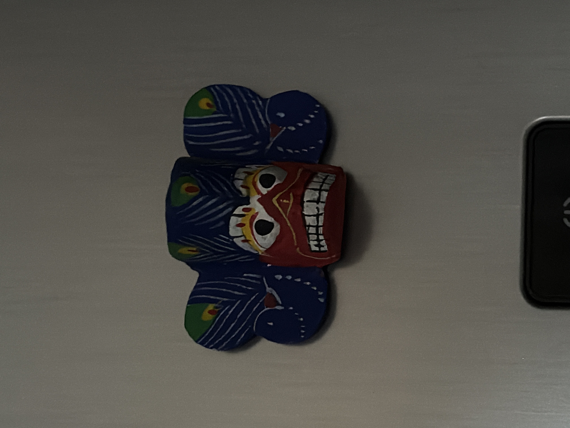 A colorful, carved wooden mask is affixed to a metallic surface. The mask is predominantly blue with red and white accents, featuring painted peacock feather-like designs. It has large, white eyes with black pupils, a red face with a wide, toothy grin, and blue, wing-like extensions on either side of the head. The surface appears to be a refrigerator door in a Moscow apartment.