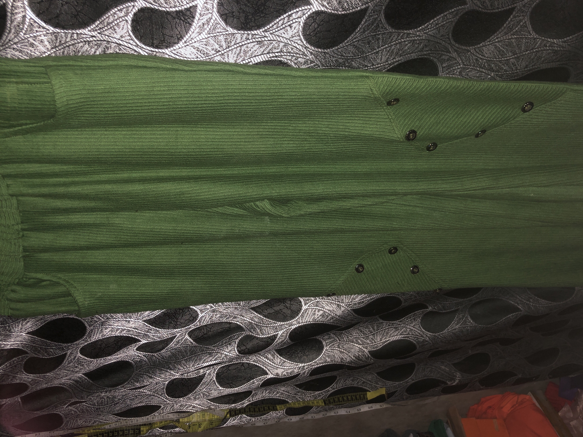 A close-up, medium shot shows the lower half of a pair of green corduroy pants. The pants are ribbed vertically and have two pockets on the front. There are decorative button details on the lower portion of the pants, arranged in a V-shape on each leg. The pants are hanging against a patterned curtain that features black and white swirling designs. A yellow measuring tape is visible on the left side of the frame, partially obscured by the curtain. The background is dark and appears to be an indoor setting. No people are visible in the image. The scene is likely in a retail or personal space in Ado, Nigeria, given the presence of a measuring tape and the typical materials seen in such environments.