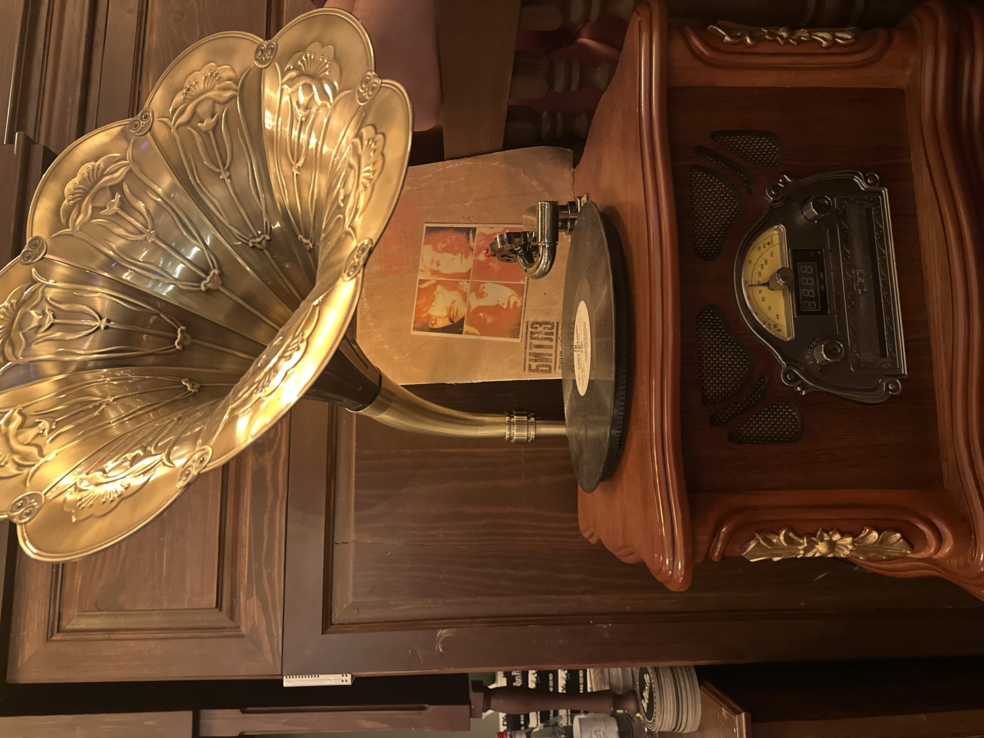 A top-down view shows an antique phonograph with a large, ornate gold horn. A vinyl record sits on the turntable, next to a record cover with four faces printed on it in a 2x2 grid. The phonograph's base is made of wood with a radio interface. Below, storage cabinets are visible, including a white plastic item. The image appears to be taken indoors, but the location in Yerevan, Armenia is unknown from the image alone.