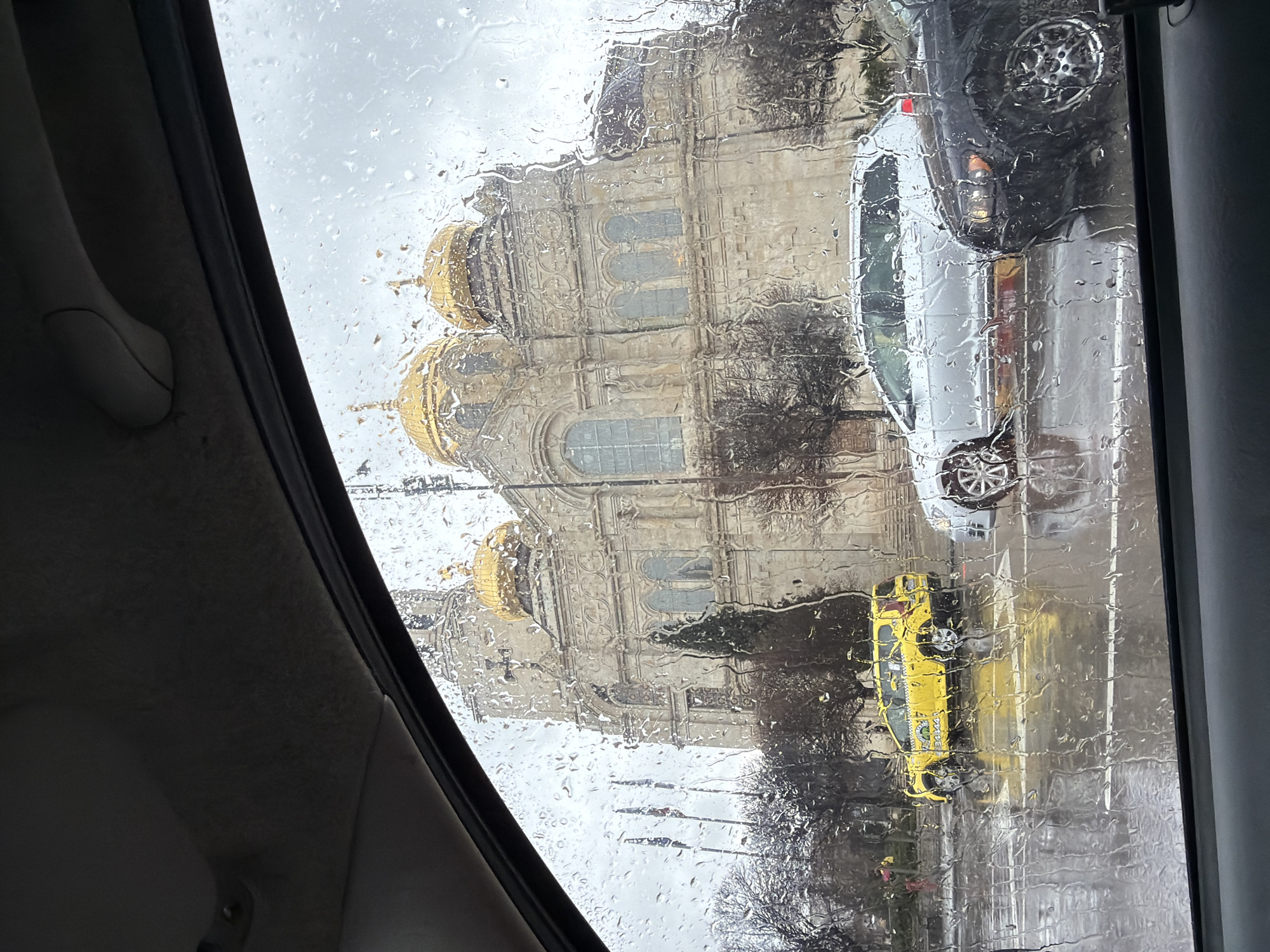 The image, taken from inside a vehicle on a rainy day, shows a view of the Cathedral of the Assumption of the Virgin in Varna, Bulgaria. A yellow taxi cab and a white van are visible on the street outside the window. Rain obscures the view of the building.