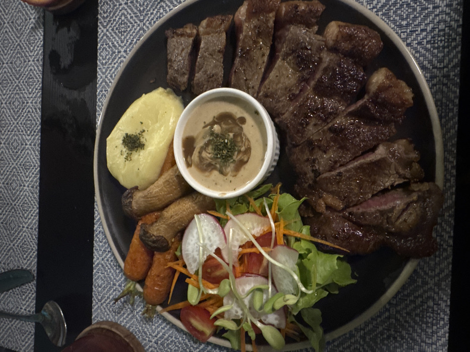 A dark, circular dinner plate is centered, presenting a prepared meal in a top-down, close-up view, consistent with a dining establishment in Ban Ko Li Pe, Thailand. The plate holds a sliced, grilled steak, appearing medium-rare to medium, distributed across the right and lower right portions. A small, white, fluted ramekin in the center contains a creamy, light brown sauce, possibly mushroom gravy, garnished with chopped green herbs. Adjacent to the sauce, in the upper right quadrant, is a portion of smooth, pale yellow mashed potatoes, topped with dried green herbs. The upper left of the plate contains several whole, roasted orange carrots and two large, elongated, sautéed mushrooms, light brown in color. A fresh green salad occupies the mid-left, consisting of green lettuce, sliced red cherry tomatoes, thin strips of orange carrot, and thin slices of white radish, topped with green sprouts. The plate rests on a placemat featuring a repeating blue and white geometric pattern. A dark, possibly black, rectangular table runner is visible horizontally across the upper background. A metallic utensil, likely a spoon, and the rim of a dark-colored cup or glass are partially visible in the upper left background. No individuals or active interactions are depicted.