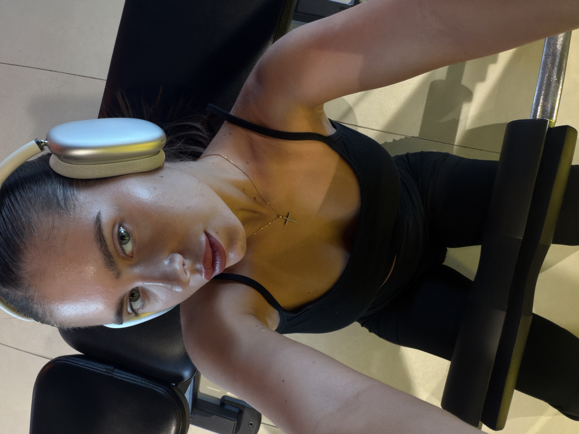 This image depicts a young adult female captured in a self-portrait within a gym environment in Krasnoyarsk, Russia. She has dark hair pulled back and wears light-colored over-ear headphones. Her attire consists of a black spaghetti-strap top and black leggings. A thin gold-colored chain necklace with a small cross pendant is visible on her chest. Her lips are adorned with red lipstick, and her skin shows a luminous sheen, possibly from physical activity. She is positioned on or near gym equipment, with a prominent black padded bar in the immediate foreground. In the background, sections of a black padded bench and a metallic, textured bar, consistent with gym apparatus, are visible. The floor is light-colored tile. The scene is illuminated by artificial light, creating distinct shadows.