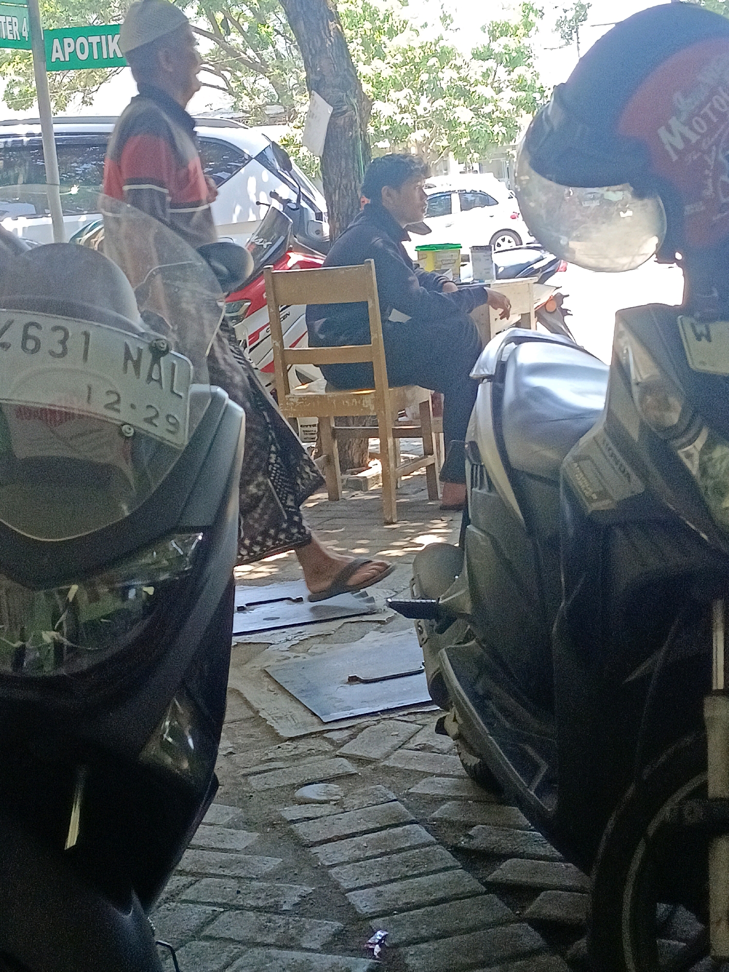 In Jimbaran Kulon, Indonesia, an outdoor scene depicts two individuals and several motorcycles. In the foreground, a dark-colored motorcycle is partially visible, with its front fender and wheel in the lower left portion of the frame. To the right, another motorcycle, predominantly dark gray with metallic accents, is seen from a similar low angle, showing its side panel and a portion of its wheel.
Behind these motorcycles, a wooden chair is occupied by a man wearing a dark long-sleeved shirt and dark pants. He is seated and facing left, with his left arm resting on the chair back. To the left of this man, a person wearing a light-colored cap and a jacket with red and gray stripes is standing. This individual is partially obscured by the motorcycle in the foreground.
In the background, a white car is parked, and trees with dense foliage provide shade. A sign with the text "APOTIK" (pharmacy) is visible on a pole, suggesting the location is near a commercial area. The ground is paved with interlocking brick pavers, with some metal sheets laid out in patches. The overall lighting indicates a bright, sunny day.
