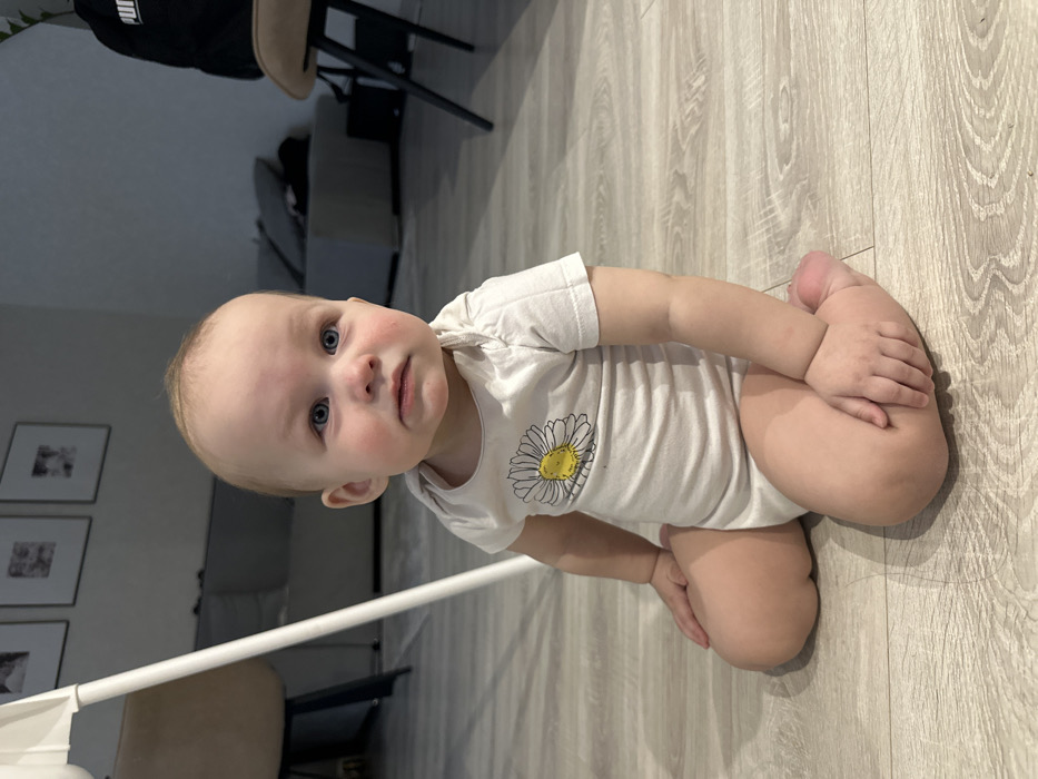 An infant is seated upright on a light-colored wood-grain laminate floor in a residential interior located in Chaikovsky, Russia. The infant, estimated to be between 6 and 18 months old, has fair skin, light hair, and blue eyes, looking forward with a neutral expression. The infant wears a short-sleeved white onesie featuring a yellow and white daisy illustration on the chest. To the left of the infant, the white support leg of a highchair is partially visible. In the background, a light-colored sofa or sectional is positioned against a light-colored wall, which displays three framed pictures. To the right, a chair with dark legs and a light-colored upholstered seat is partially visible, with a black garment marked with a "PUMA" logo draped over it, and a dark object situated behind the chair.