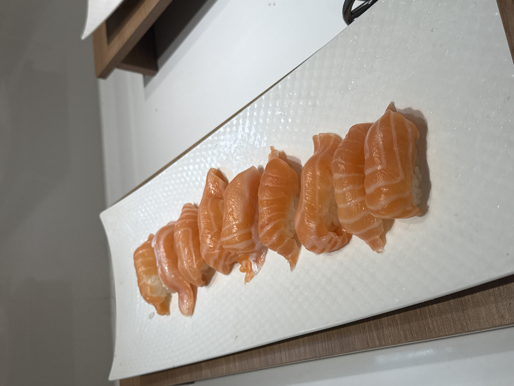 A close-up, high-angle shot shows a white rectangular plate filled with ten pieces of salmon nigiri sushi. The sushi is arranged in a line, with the salmon pieces facing the viewer and slightly overlapping each other. The salmon is a vibrant orange color with visible white fat marbling. Each piece of sushi rests on a small bed of white rice. The plate has a textured surface. The background consists of a neutral gray wall and a wooden surface. The scene is well-lit, highlighting the freshness of the sushi. The location in Nha Trang Ward, Vietnam, is not visually identifiable from the image content.