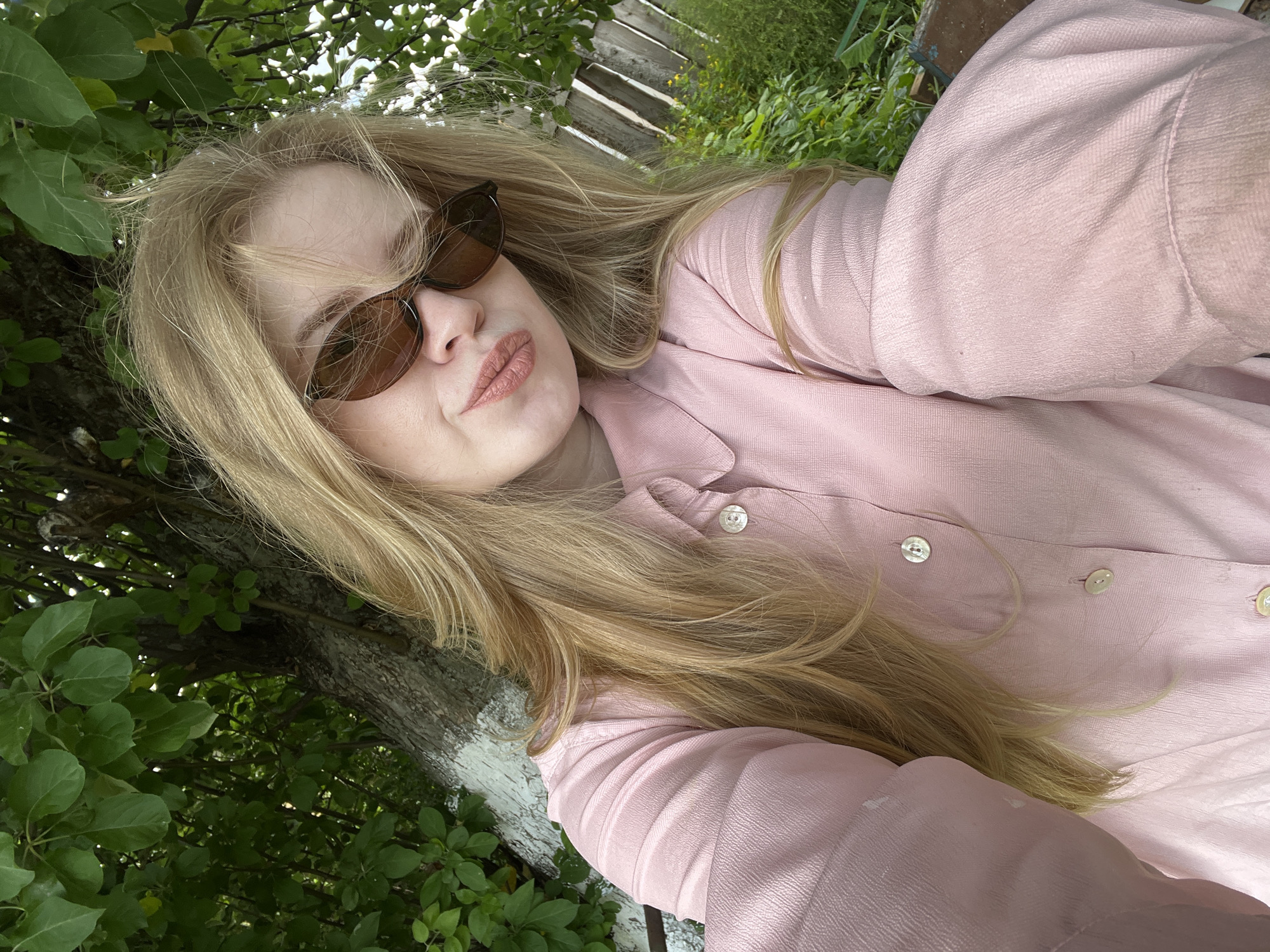 A young woman with long blonde hair and sunglasses takes a selfie outdoors. She is wearing a pink collared shirt with buttons. She is sitting in front of a tree with green leaves and a wooden fence in the background. The location appears to be a park or backyard. There is no visual information to suggest this is in Chaikovsky, Russia.