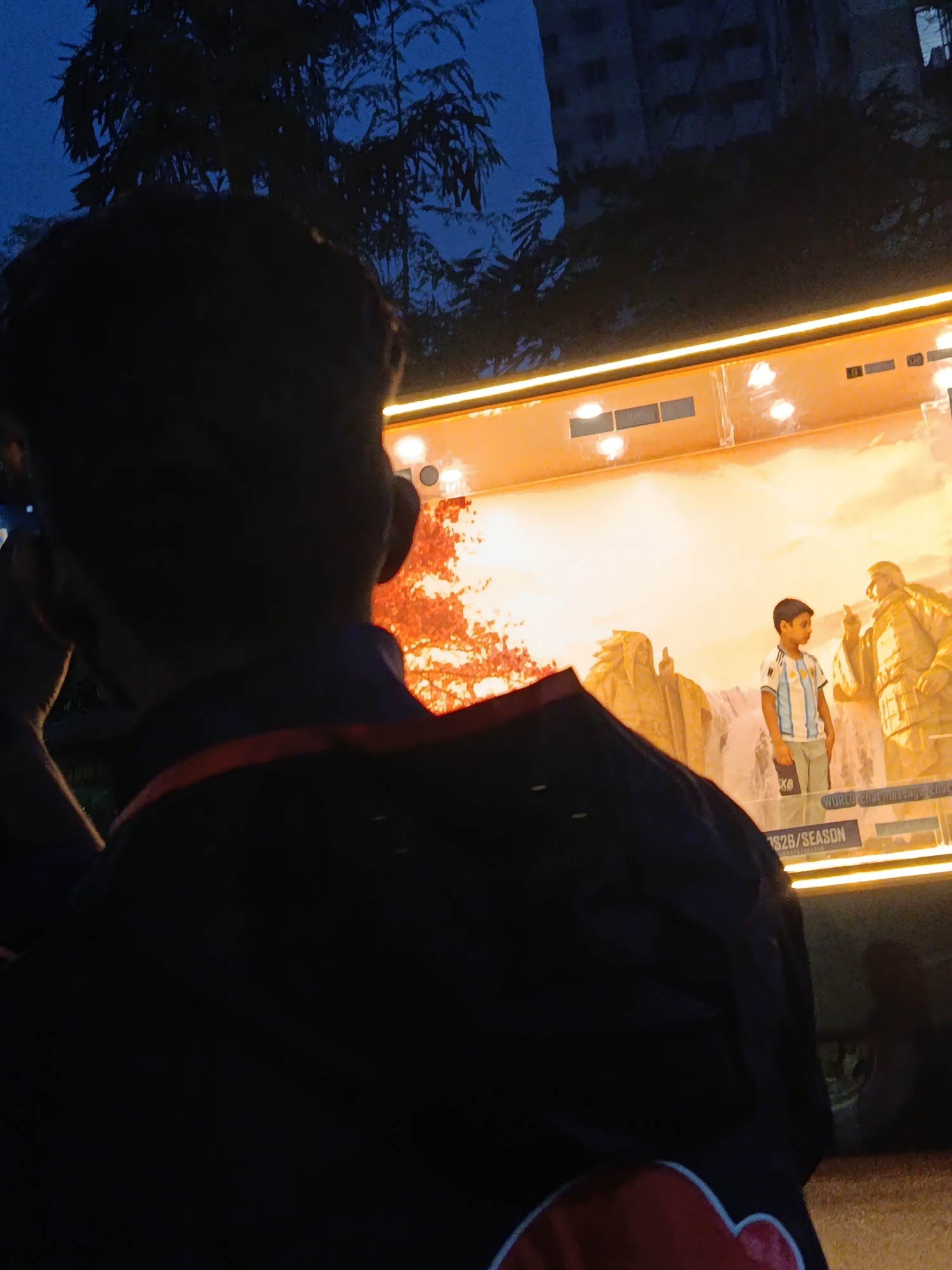 A person is viewed from behind, facing a brightly lit display with an image of a boy in a soccer jersey interacting with a large, golden statue. The display is set against a backdrop of a rocky landscape with waterfalls and foliage. Text reading "WORLD chat message" and "FS2B / SEASON" is visible below the image. The scene appears to be outdoors at dusk, with trees and a building visible in the dark background. The person in the foreground is wearing a dark jacket with red trim. The location is identified as Dhaka, Bangladesh.