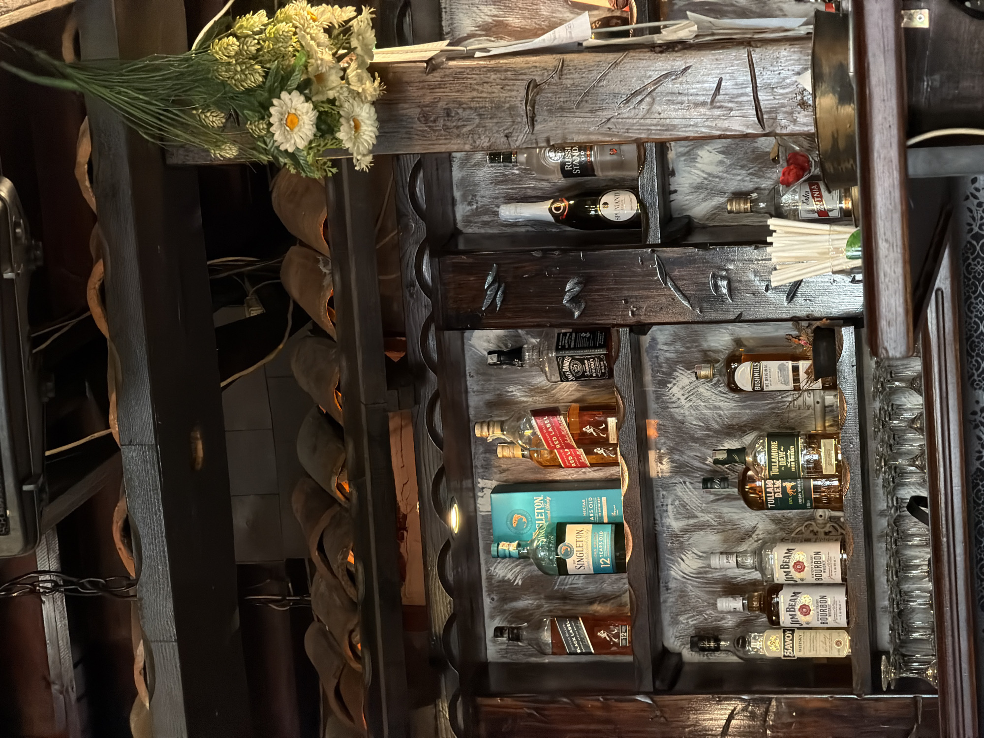 This image depicts the interior of a bar or restaurant located in Varna, Bulgaria. The scene focuses on a rustic-style liquor display featuring dark, distressed wooden shelving units against a mottled grey and white back paneling.
The upper portion of the image reveals dark wooden beams, a black metal chain, and an overhead appliance. Below these, multiple rows of terracotta-like, half-round decorative tiles are present, each exhibiting a warm orange glow from internal lighting.
On the top visible shelf, from left to right, are a bottle of Johnnie Walker Black Label 12-Year-Old Scotch Whisky, a bottle of The Singleton 12-Year-Old Single Malt Scotch Whisky accompanied by its blue packaging box, two bottles of Johnnie Walker Red Label Scotch Whisky, and a partially visible bottle of Jack Daniel's Tennessee Whiskey.
The middle section of the shelving, divided by vertical wooden uprights, includes a bottle of Jack Daniel's Tennessee Whiskey, a bottle of Spumante Brut sparkling wine with a silver foil top, and a bottle of Russian Standard Vodka. Silver-colored, leaf-shaped metallic decorations are affixed to the wooden elements. A cluster of artificial white daisies with yellow centers and green hop-like foliage hangs from an overhead beam on the right side.
The bottom shelf, positioned above a bar counter, displays a selection of spirits: a bottle of Savoy Whisky, two identical bottles of Jim Beam Bourbon Whiskey, two bottles of Tullamore D.E.W. Irish Whiskey, and a partially visible bottle of Bushmills Original Irish Whiskey.
Below this shelf, numerous inverted stemmed glasses are neatly arranged on a rack. On the dark wooden bar counter in the foreground, specifically on the right, a clear glass container holds numerous white cocktail straws. Adjacent to the straws, a green lime wedge rests on the counter surface. A small, clear bowl contains red berries, and a golden metallic bowl or tray is positioned nearby. A bottle labeled "Extra Zhitnitsa" is visible behind the straws. No individuals are present in the depicted scene.