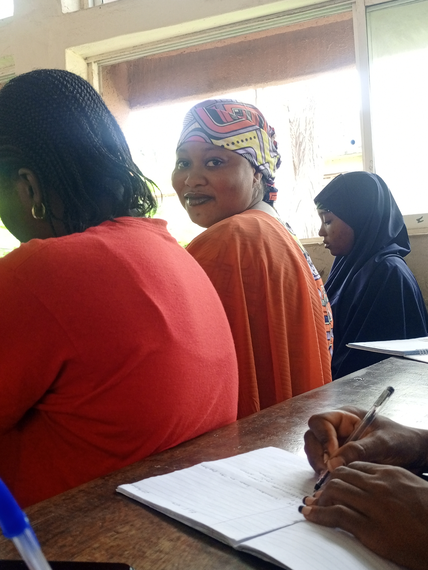 The image shows a classroom setting with several women sitting at desks. One woman in the center is looking towards the camera, wearing an orange top and a colorful headscarf. To her right, a woman is wearing a black hijab. To the left, the back of another woman wearing a red top is visible. In the foreground, a person's hands are writing in a notebook with a pen. A blue pen is also visible on the desk. The location is in the city of Unknown, Unknown.
