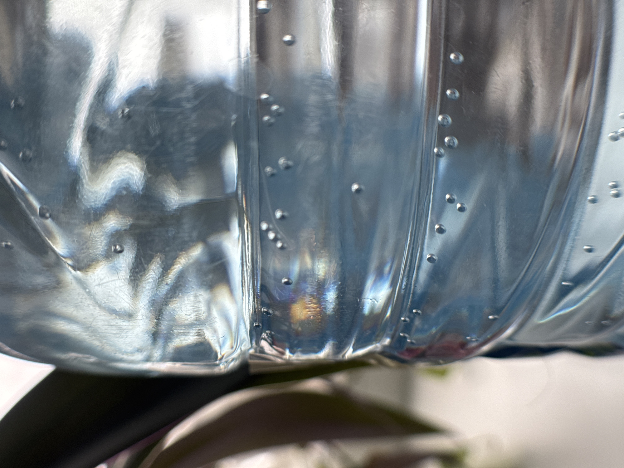 The image shows a close-up view of the lower portion of a clear plastic bottle, likely filled with water. The bottle has a ribbed or contoured design, with numerous small air bubbles clinging to the inner surface. The plastic is slightly distorted, creating a wavy effect and refracting light. The bottom of the bottle rests on a dark, slightly blurred surface, possibly a table or stand. In the background, there is a blurry glimpse of vegetation. There is no indication of location or inhabitants.