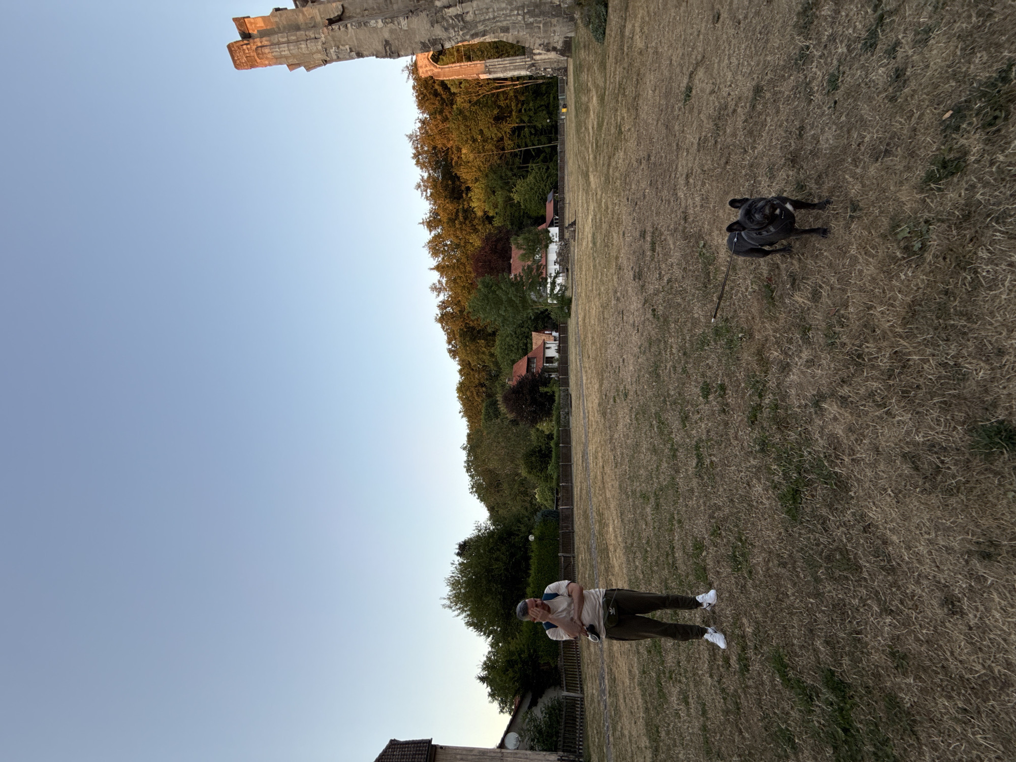 In Walkenried, Germany, an outdoor scene unfolds under a clear blue sky with the setting sun casting a warm glow on the trees in the background. In the foreground, a man stands in a dry, grassy field, his right hand near his face, appearing contemplative. He is wearing a light-colored t-shirt, dark pants, white sneakers, and a fanny pack. A French bulldog on a leash stands to his right, facing the camera. To the left of the man, a portion of a building is visible, and to the right, the ruins of an ancient stone structure rise, its weathered stone illuminated by the sunlight. In the middle ground, several houses with red-tiled roofs are nestled amongst the trees, forming a small village.
