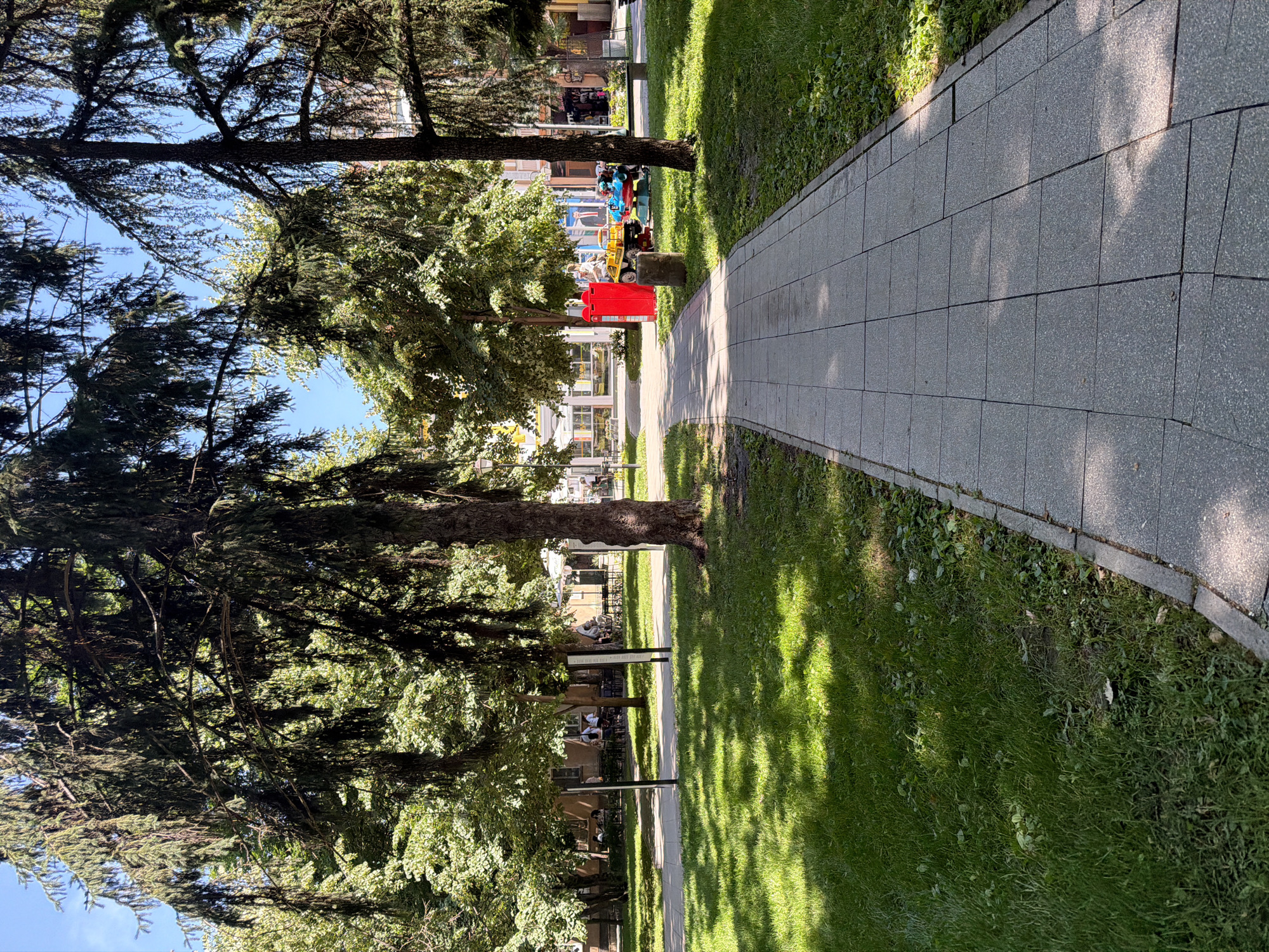 The image shows a paved walkway running alongside a grassy area with trees. The walkway is made of gray, square-shaped paving stones and is bathed in sunlight, casting dappled shadows from the trees. To the left of the walkway, lush green grass is visible, with some patches of clover and other low-lying plants. Several trees, with dark bark and full green foliage, line the edge of the grass.
In the background, a building with multiple windows and a light-colored facade can be seen, suggesting an urban or park setting. A red trash receptacle stands near the walkway, and behind it, a colorful children's ride with various amusement park elements is visible, with people, including children, appearing to be interacting with it. Further down the walkway, a few people are seated at tables under a white umbrella, possibly at an outdoor cafe.
The scene appears to be set during the day, with bright sunlight indicating clear weather. The shadows cast by the trees suggest it could be mid-morning or afternoon. The presence of trees, grass, a walkway, and people enjoying leisure activities implies a park, public garden, or a well-maintained urban outdoor space. There is no visible text in the image.
