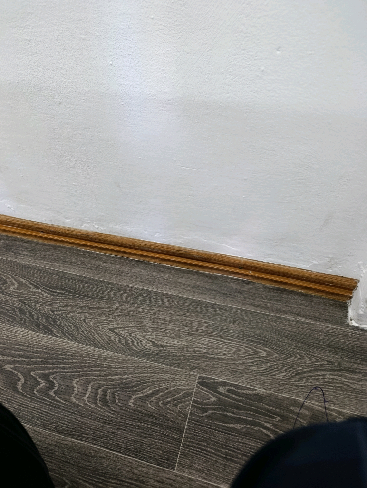 The image shows a close-up view of a wall and floor in what appears to be an interior space. The wall is painted white and has a textured surface with some imperfections. A wooden baseboard runs along the bottom of the wall, transitioning to a dark grey wood-look laminate flooring. The floor planks have a distinct grain pattern. The scene is devoid of any people or specific actions. The lighting suggests an indoor environment. There are no identifying landmarks or details that would allow for pinpointing a location in Kostanay, Kazakhstan.