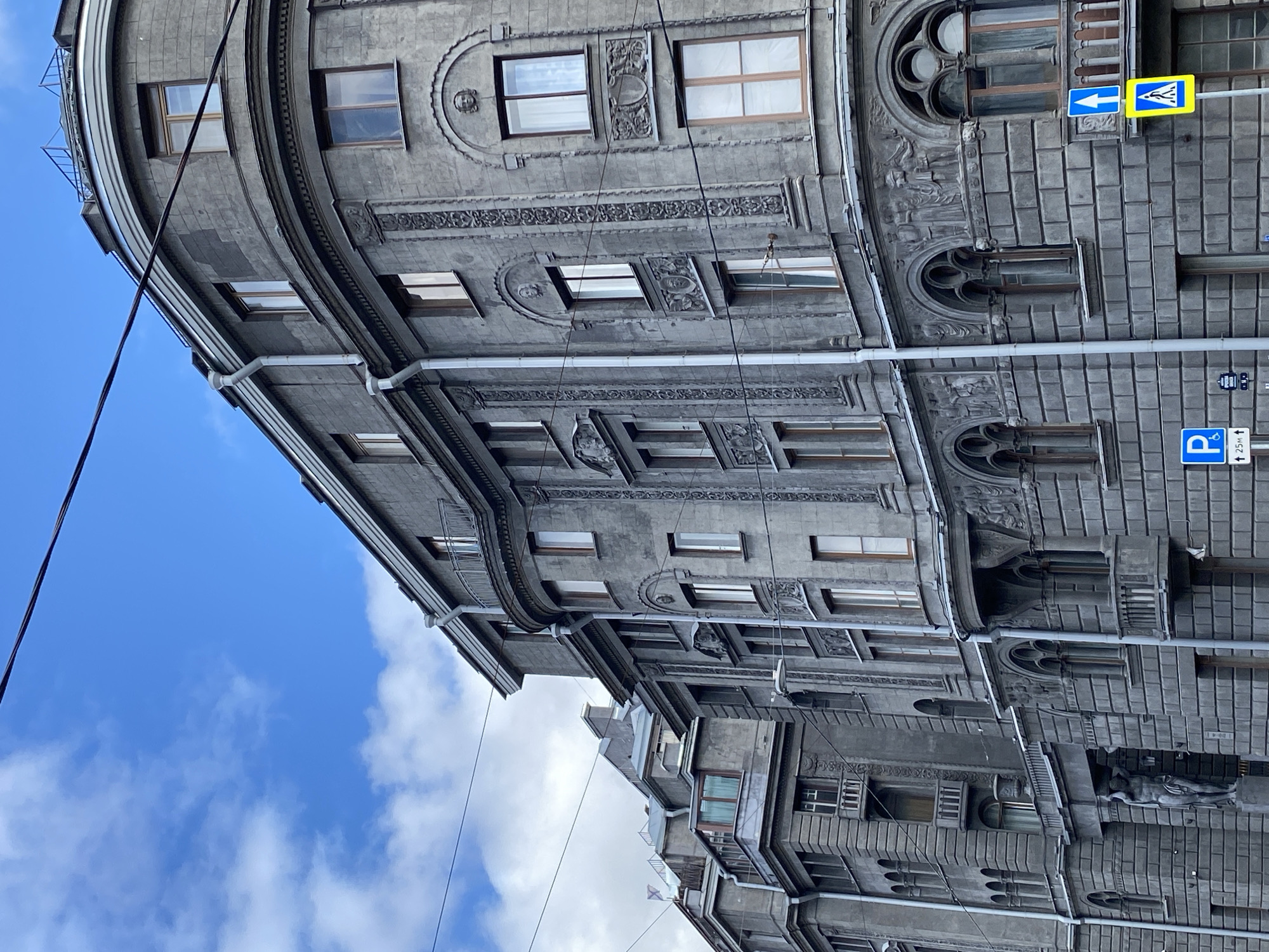 This is a medium-angle, full shot of the facade of an old building in Saint Petersburg, Russia, with a cloudy sky above. The building is constructed of grey stone and features ornate architectural details such as decorative carvings, arched windows, and balconies. A statue of a male figure can be seen in the lower left portion of the frame, partially obscured by the building's architecture. Overhead, electrical wires stretch across the sky, and a street sign with a blue arrow indicating direction and a yellow sign with a pedestrian symbol are visible on the right side of the image. The building appears to be multi-story, with numerous windows and distinct architectural sections.