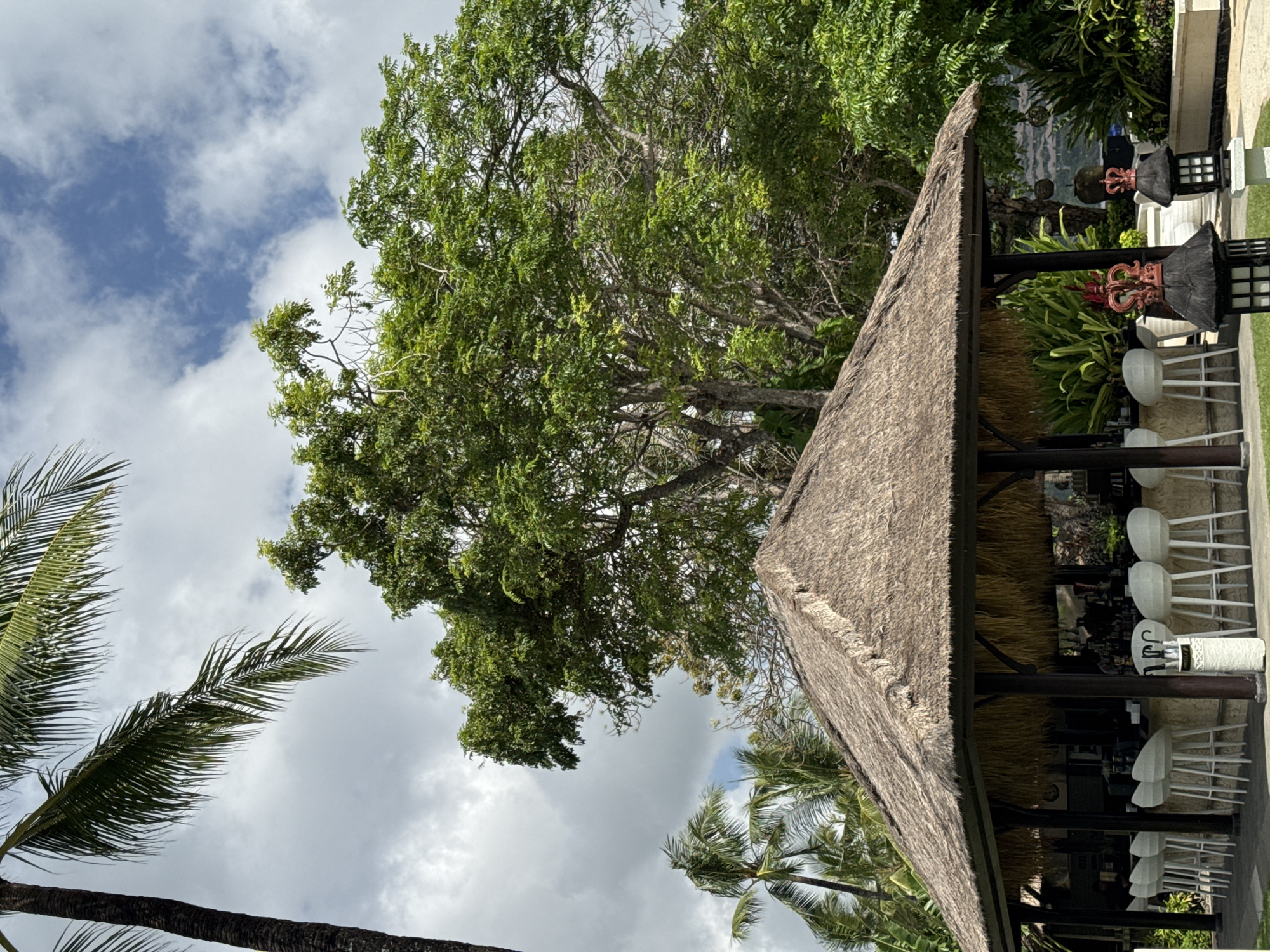 The image captures a scene in Kuta Selatan, Indonesia, likely a resort or outdoor dining area. The primary focus is a thatched-roof structure, indicative of tropical architecture, likely housing a bar or restaurant. A row of white bar stools lines one side of the structure. The background shows dense tropical foliage, including various trees. The sky overhead features a mix of blue and white clouds, suggesting a partly cloudy day. Palm trees are also visible, enhancing the tropical ambience. The photograph is taken from a low angle, emphasizing the height of the thatched roof and the surrounding trees. In the background there are decorative elements on the building.