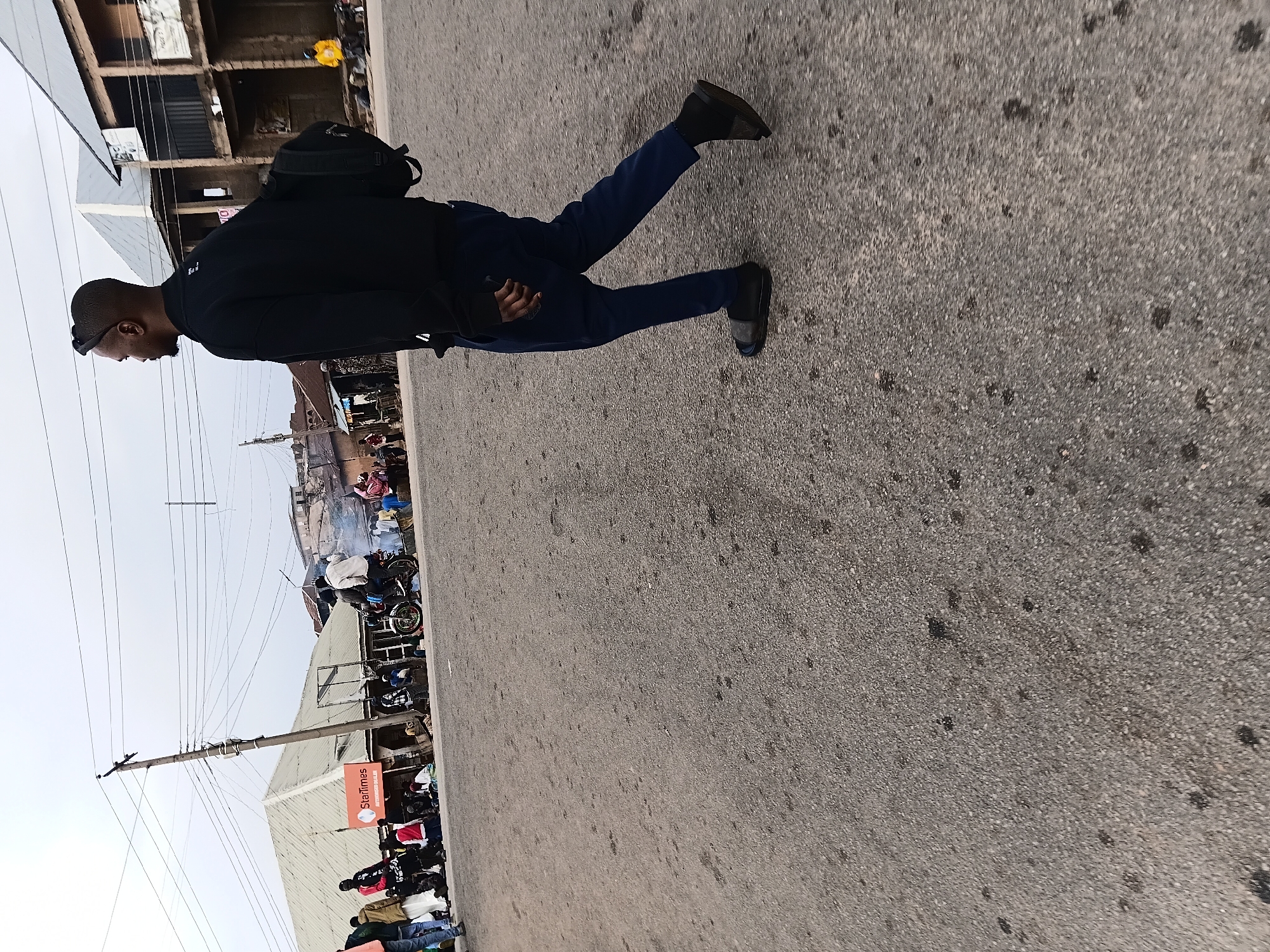 A man with a shaved head, wearing a dark long-sleeved shirt, blue pants, and dark shoes, is walking along a paved road in Gwong, Nigeria. He carries a dark backpack and appears to be holding a phone in his left hand.
In the background, there are buildings, power lines, and a crowd of people gathered near a "StarTimes" advertising sign. Some motorcycles are parked nearby. The weather appears to be overcast.
