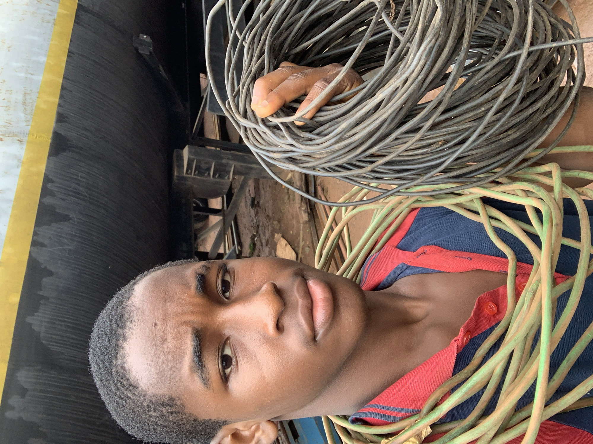 A young African male with dark skin, short, wavy hair, and a serious expression is in the foreground of the image. He is wearing a blue polo shirt with red trim and a red collar, with green and brown cables draped around his neck and shoulders. He is holding a large coil of thick, grey wire in his right hand. Behind him, a large, dark, industrial structure with a yellow stripe runs along the top. The ground appears to be dirt with some debris. The image was taken in Nkpor, Nigeria.
