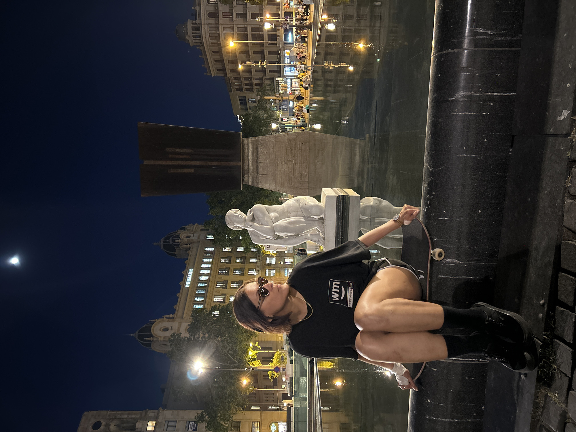 A nighttime cityscape features a person seated with a skateboard beside a fountain or reflecting pool, positioned at the edge of a street or square. The person is wearing sunglasses, a dark T-shirt with graphics, short shorts, black socks, and dark shoes, leaning back with one hand resting on the skateboard. Behind the person, a modernist sculpture of two figures lies on a pedestal, next to a large, dark rectangular sculpture. The scene is illuminated by city lights, creating reflections on the water. In the background, several multistory buildings with lighted windows line the square, including a building with the name "PRIMARK" visible, pinpointing the location as Plaça de Catalunya in Barcelona, Spain. The night sky above is dark, with a visible moon.