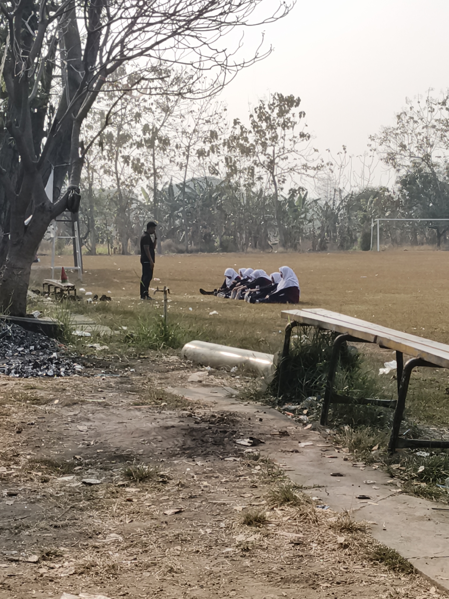 A group of five individuals, all appearing to be female and wearing hijabs and dark clothing, are sitting on a grassy field. They are positioned in a line, facing towards the right of the frame. To the left of the group, a male figure dressed in a dark shirt and pants stands upright, facing towards the left, with his hand near his face. Behind the seated group, a soccer goal is visible in the distance. The field is covered in dry grass and scattered debris. In the foreground, a concrete pathway is present, also littered with debris. A long, weathered wooden bench with dark metal legs is situated in the middle ground, to the right of the frame. The overall environment suggests an outdoor athletic field with areas of untidiness.