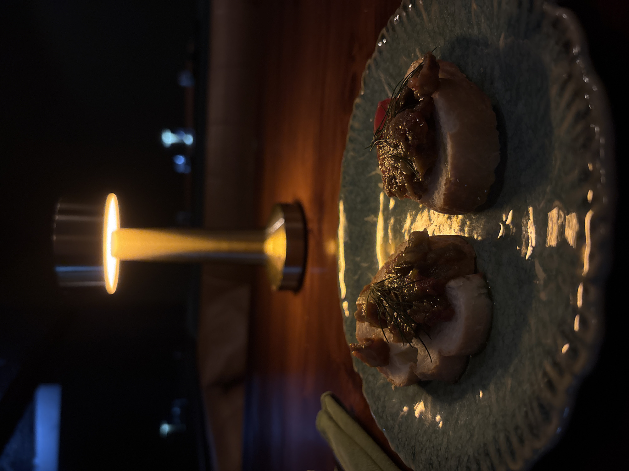 The image shows a dimly lit scene in Kuta Selatan, Indonesia. Two small, open-faced sandwiches sit on a light blue plate with a decorative, scalloped edge. Each sandwich appears to have a circular slice of white bread topped with a dark brown mixture, garnished with green sprigs, and one has a small piece of red. A small lamp with a cylindrical shade is visible, casting a yellow glow on the sandwiches and the wooden table. The background is dark. A green item is in the lower foreground.