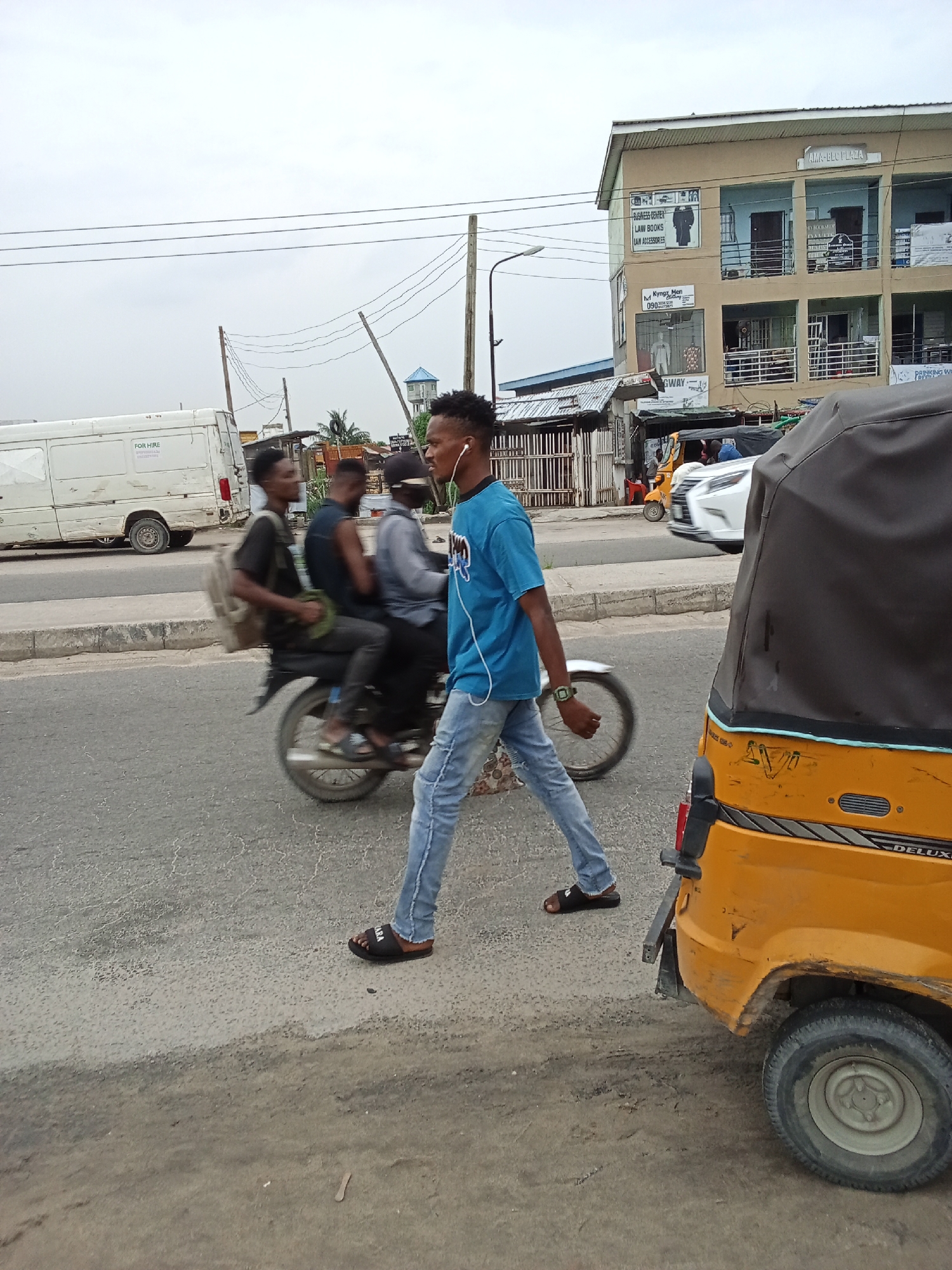 The image depicts a street scene in Ado, Nigeria. In the foreground, a young man in a blue t-shirt, light jeans, and sandals is walking across the street, listening to headphones. To his right is a yellow "DELUX" tricycle taxi. Behind him, a motorcycle carries three passengers, moving from left to right. A white van with "FOR HIRE" text is parked on the left side of the street. Across the street, partially visible, is a multi-story building with signage that includes "BUSINESS CENTER," "LAW BOOKS," "LAW ACCESSORIES," "Kyngz Men" and "AMA-BLC PLAZA". Overhead, utility poles and wires run across the sky. The weather appears overcast.