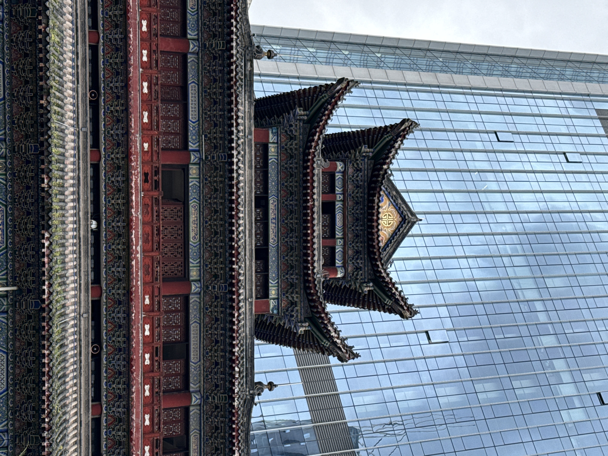 The image shows a traditional Chinese building juxtaposed against a modern glass skyscraper in the Yuzhong District of Chongqing, China. The ornate building is detailed with red, blue, and green ornamentation. The skyscraper is mostly glass.