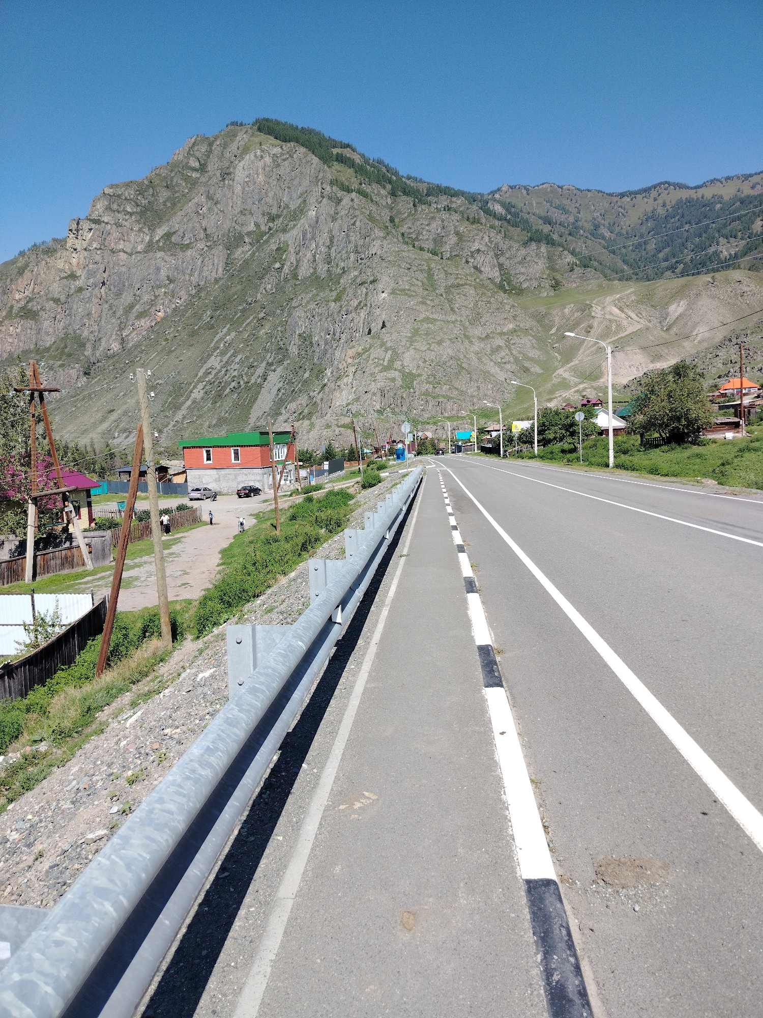 The image shows a scenic view of a road passing through a mountainous, rural area. A paved road with white lane markings curves to the right in the foreground, with a sidewalk and a metal guardrail on the left. Beyond the road, on the left side, are a few houses, some with colorful roofs, and wooden fences. A prominent, large rocky mountain dominates the background, with trees sparsely covering its slopes and the slopes of adjacent hills. The sky is a clear, bright blue. The overall environment appears to be a village in a mountainous region, possibly in Russia, given the mention of Iodro. The scene is peaceful and rural, with no discernible activity or interaction between people.