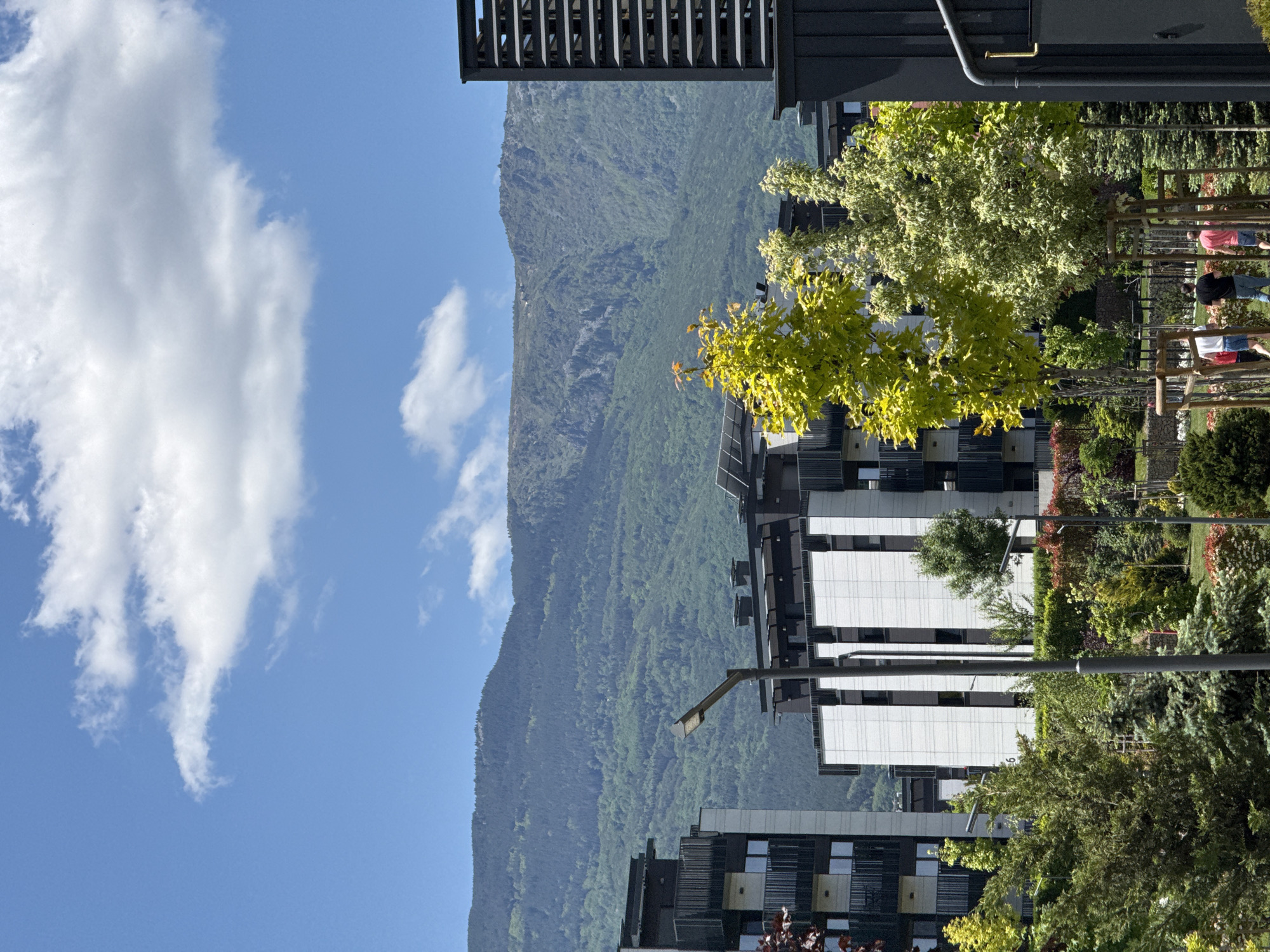 The image shows a building with a mountain range in the background, under a bright blue sky with fluffy white clouds. The architecture of the building is modern, with visible white panels and dark window frames. Several trees, varying in shades of green and yellow, are scattered in front of the building, giving the impression of a well-maintained garden or park. People are faintly visible in the park area, suggesting public access. A street light stands in the foreground, adding to the urban landscape. Due to the visible architecture, mountain range, and park setting, it may be located somewhere near Vitosha Mountain. It is possible this picture was taken in the Sofia, Bulgaria area.