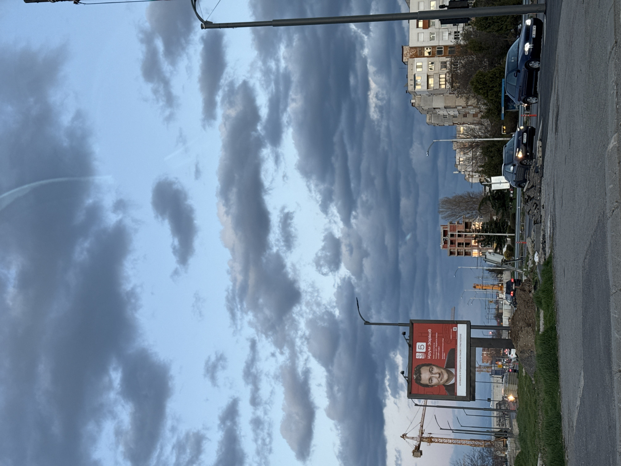 The image shows a street scene in Sofia, Bulgaria. The sky is filled with dark, heavy clouds. On the right side of the image, there is a road with cars parked on it. There is a building with many lit windows in the background. On the left side of the image, there is a billboard for a political candidate, Крум Зарков. In the distance, there is a construction site with a crane. The image is oriented vertically.
