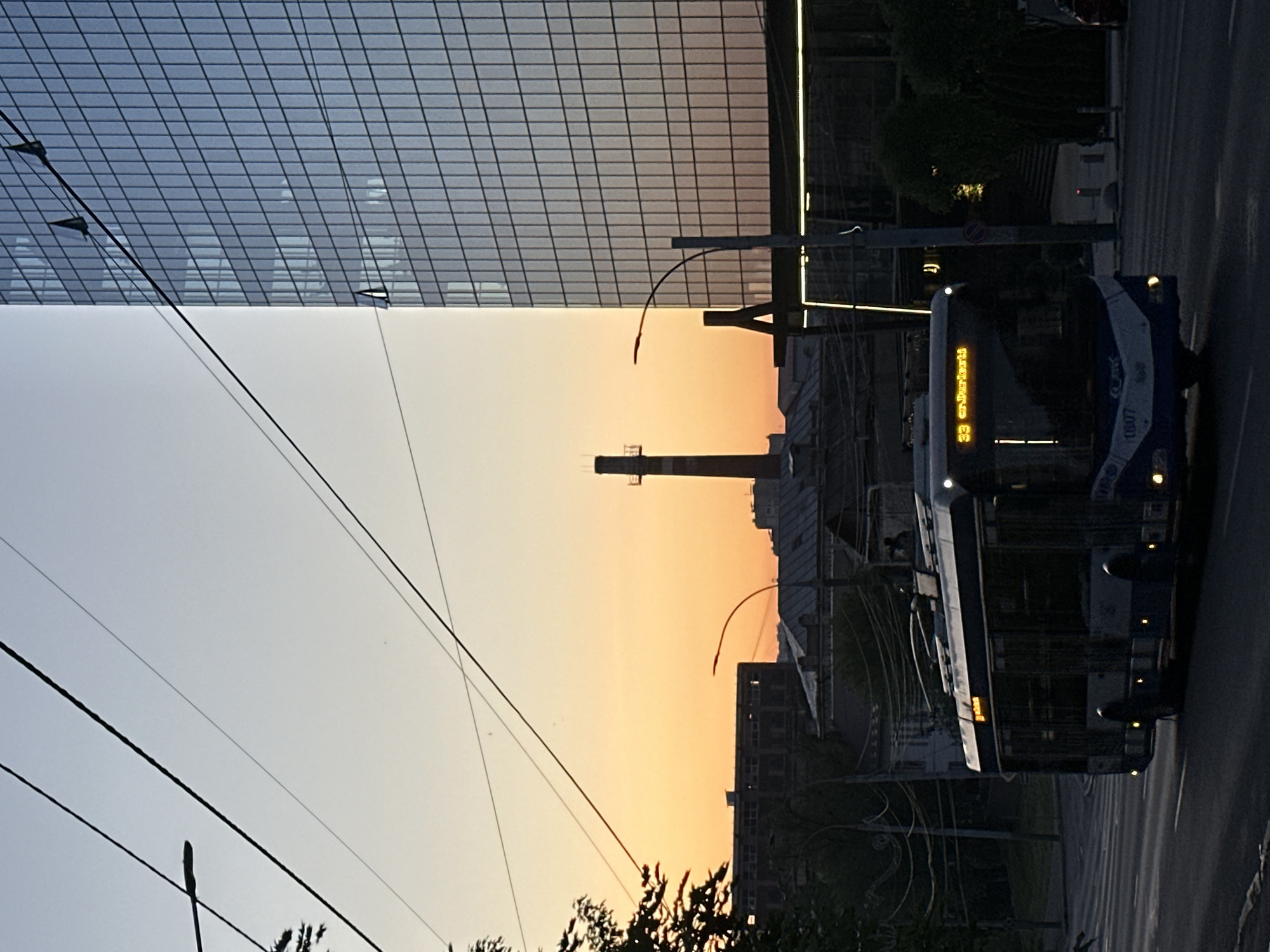 The image shows a street scene at dusk in Chișinău, Moldova. A modern glass-facade building stands on the left. Overhead power lines crisscross the sky, where the sunset creates a gradient of orange and yellow hues. In the background is a smokestack. A trolleybus, marked with the route number "33", is traveling on the street. The environment is urban, with buildings, trees, and street infrastructure present.