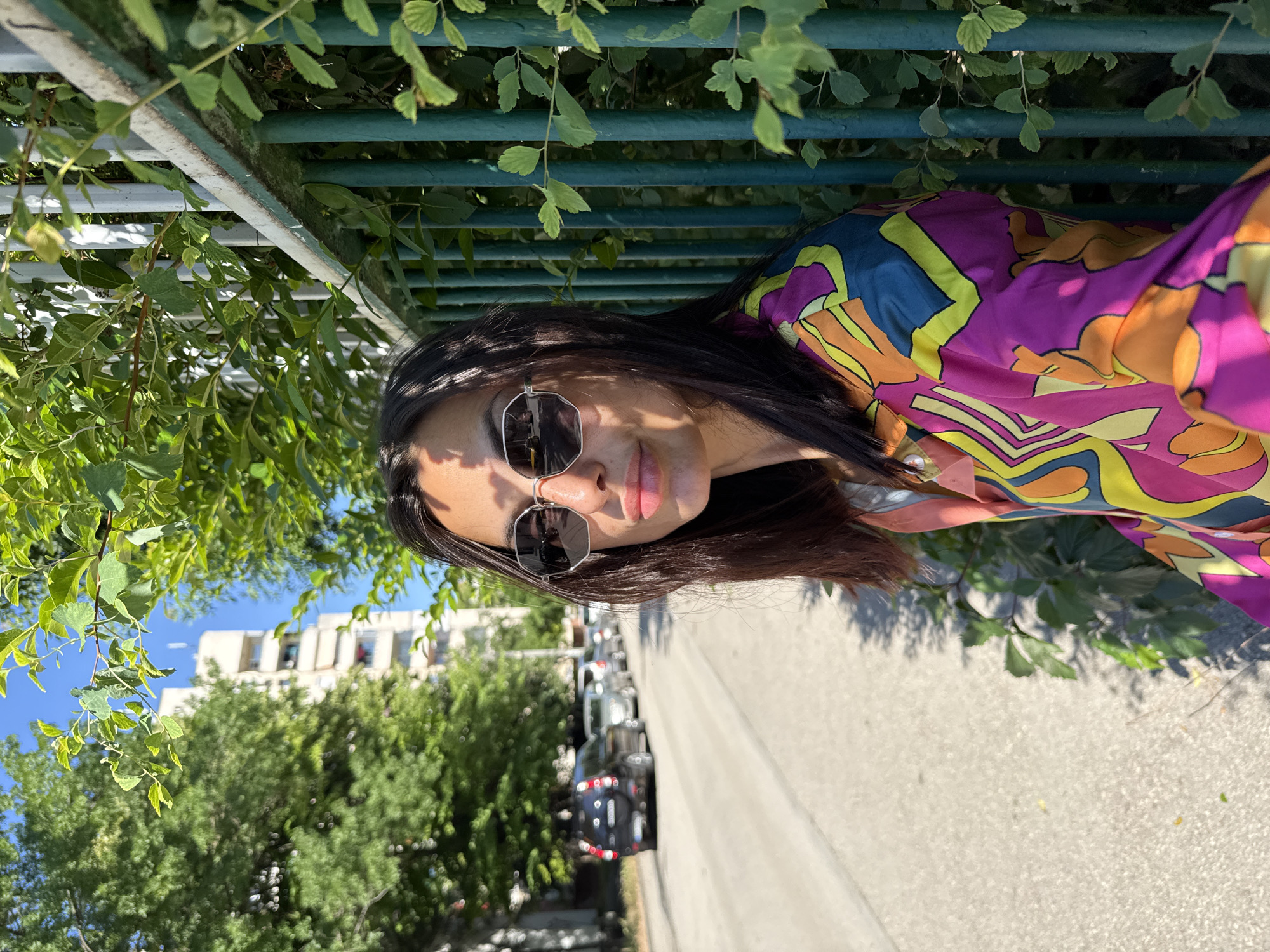 A young woman with long dark hair and sunglasses is standing on a sidewalk next to a green metal fence with lush green foliage growing around it. She is wearing a vibrant, colorful, retro-patterned shirt. Behind her, a street lined with trees and parked cars stretches into the distance, with a tall, light-colored building visible in the background. The scene appears to be in a residential area of Pleven, Bulgaria, under a clear blue sky on a sunny day.
