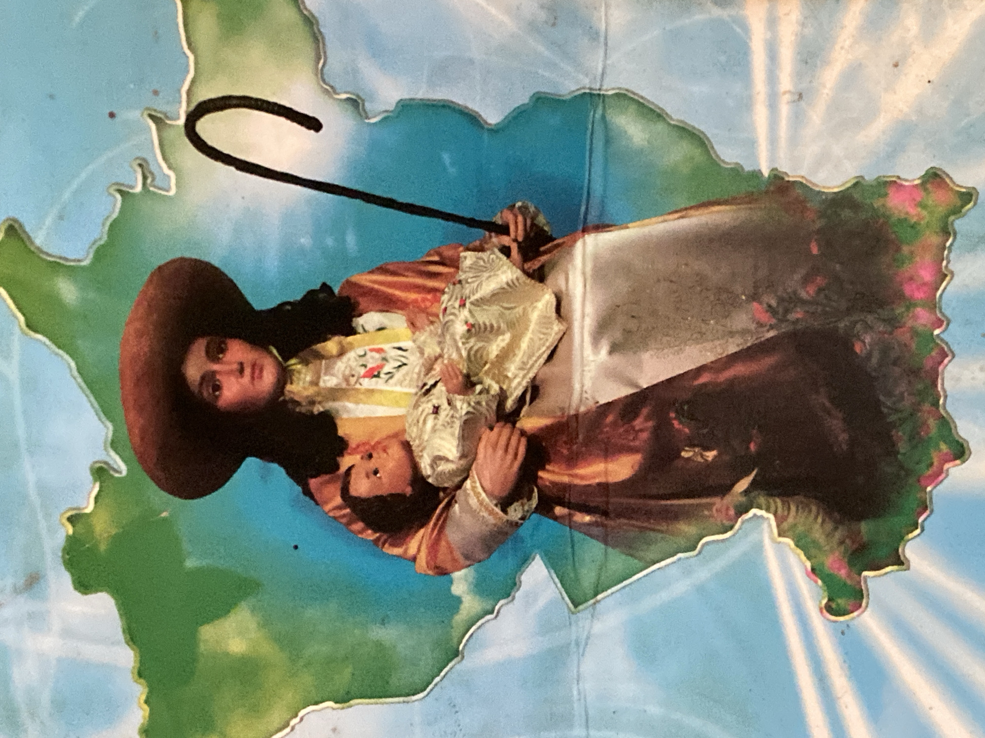 The image displays a vibrant, stylized depiction of a woman holding a child, set against a background shaped like the map of Venezuela. The woman, positioned upright and slightly rotated to the left, wears a wide-brimmed brown hat and elaborate, colorful clothing including a patterned blouse and a flowing skirt or robe in shades of gold, brown, and red, accented with floral motifs. She holds a shepherd's crook with a prominent hook in her right hand. Her gaze is directed towards the viewer, and her expression is serene. The child, dressed in ornate, light-colored garments, is cradled in her left arm, looking slightly upwards.
The figures are superimposed on an outline map of Venezuela, which features green for land and blue for water, bordered by a distinct metallic gold or silver outline. Radiant light rays emanate from behind the figures, particularly noticeable in the upper right and lower right quadrants of the background, suggesting a divine or blessed atmosphere. The composition strongly resembles a devotional image, possibly representing the Divina Pastora (Divine Shepherdess) or another Marian advocacy popular in Venezuela. No text is visible in the image. The art style suggests a piece of popular religious art or a commemorative item from a region like Tinaquillo, Venezuela, where such devotions are celebrated.