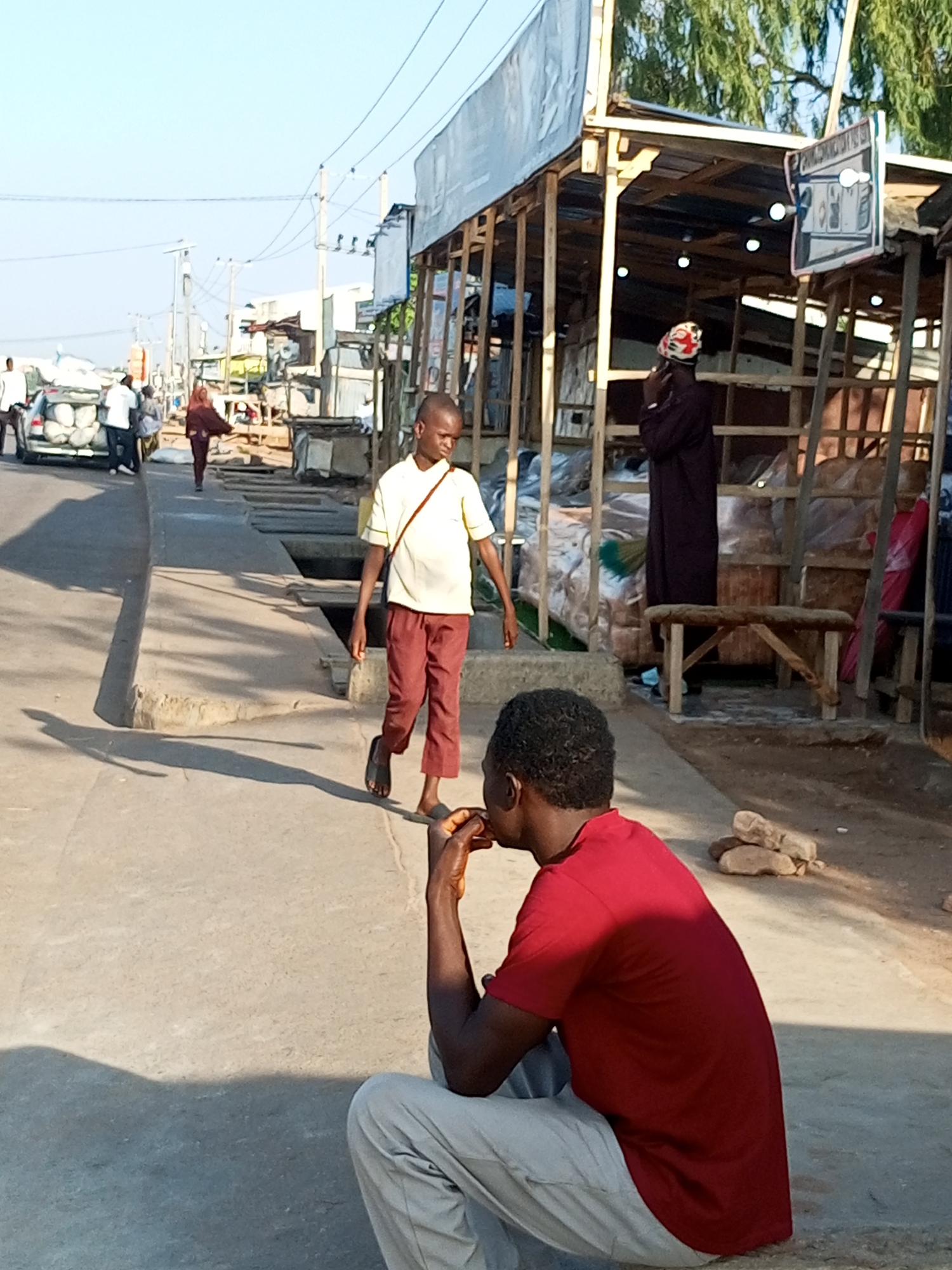 This image depicts a street scene in Garko, Nigeria. In the foreground, a person wearing a red t-shirt and grey pants sits on the ground, facing away from the camera, with their hands near their mouth. Behind them, a young man in a yellow t-shirt and maroon pants walks along a concrete pathway, casting a long shadow. To the left, a white car is parked on the side of the road, with people near it. Further back, a series of market stalls constructed with wood and corrugated metal roofing line the street. One stall displays a sign. Another stall has a person wearing a dark robe and a colorful head covering inside. The environment appears to be an outdoor market or roadside commercial area with clear blue sky overhead.