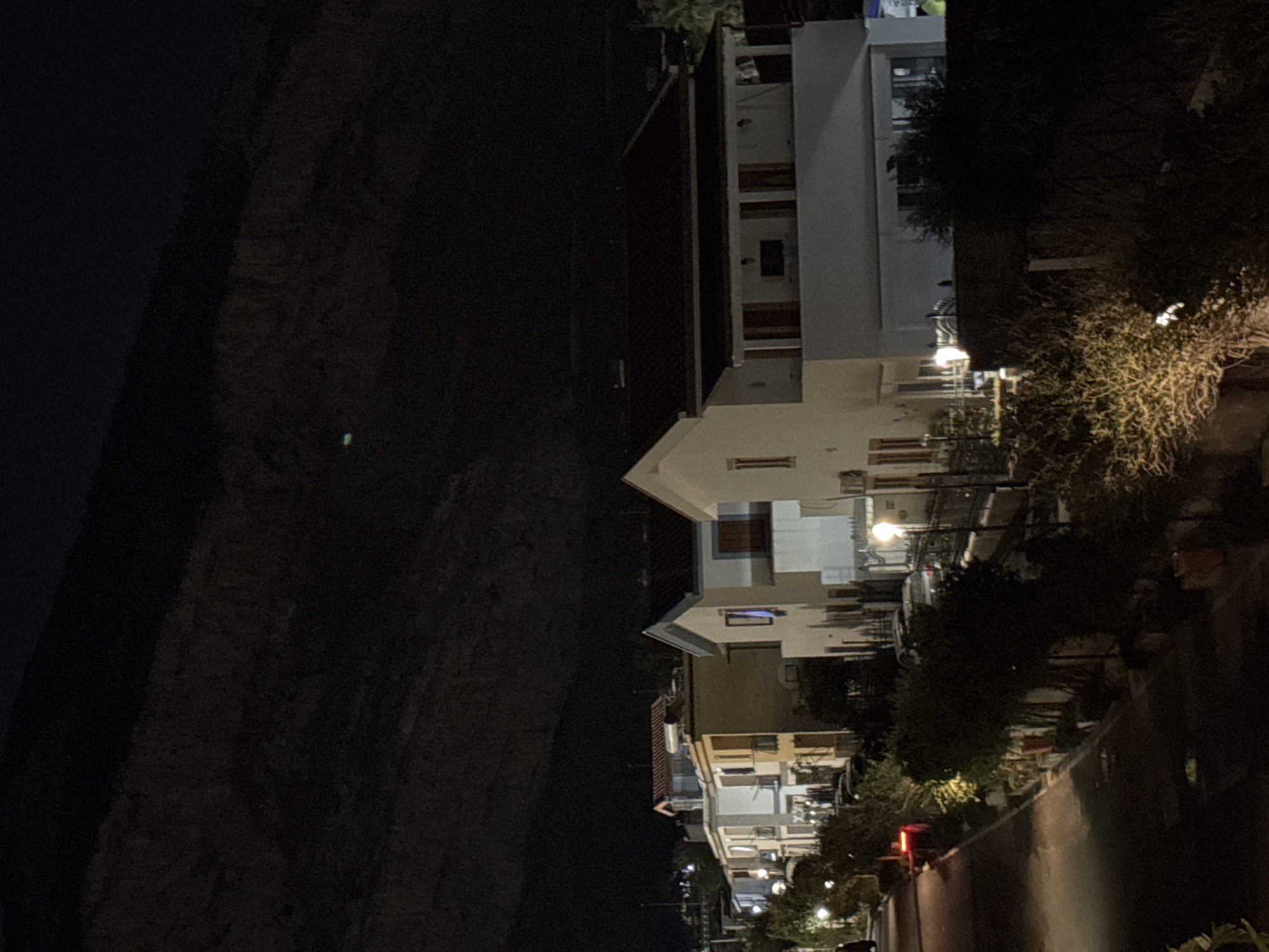 The image depicts a street scene at night in Capaci, Italy, with a row of multi-story buildings lining a road. The buildings are modern in style, with white and light-colored facades, red tiled roofs, and balconies. Some windows are illuminated from within. Trees and shrubbery are present along the side of the road, with streetlights casting a warm glow on the vegetation and paved surfaces. Several cars are parked on the street and in front of the buildings. In the background, a dark, mountainous silhouette looms against the night sky, suggesting a natural landscape bordering the residential area. The overall atmosphere is quiet and serene, characteristic of a residential neighborhood at night.
